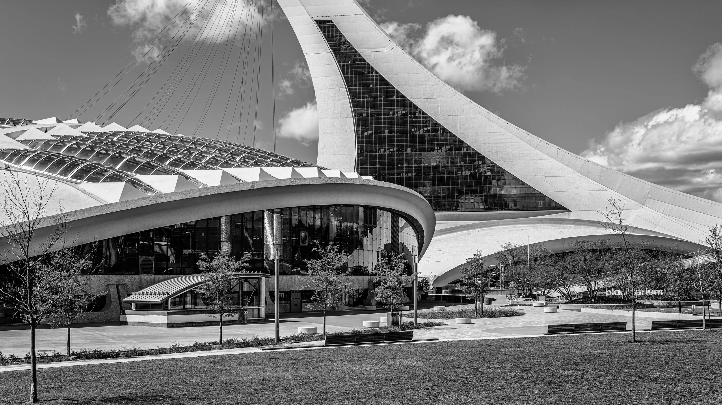 Black and white photo of a modern futuristic skyscraper with curved architecture, glass windows, and surrounding trees and benches in a park-like area.