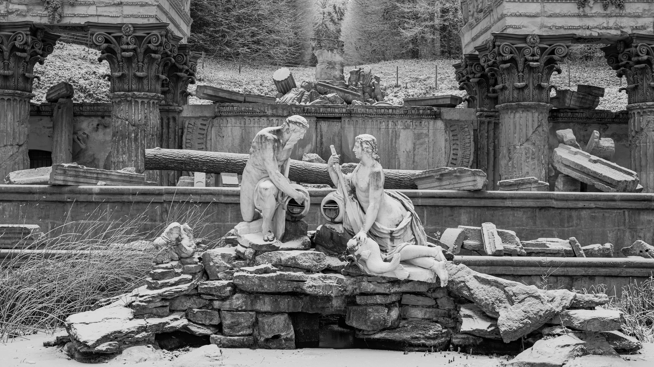 Black and white photo of ancient Greek or Roman ruins, including broken columns and statues, with snow on the ground and trees in the background.
