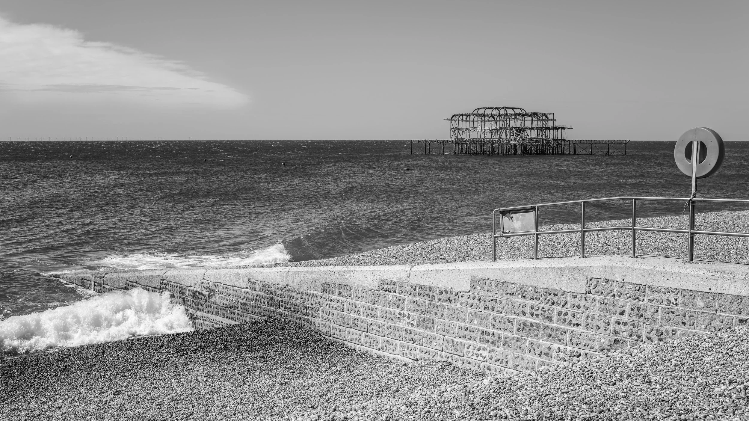 A black and white photo of a beach with a stone pier leading to the ocean, a metal railing with a lifebuoy, and a partially destroyed pier structure in the distance.