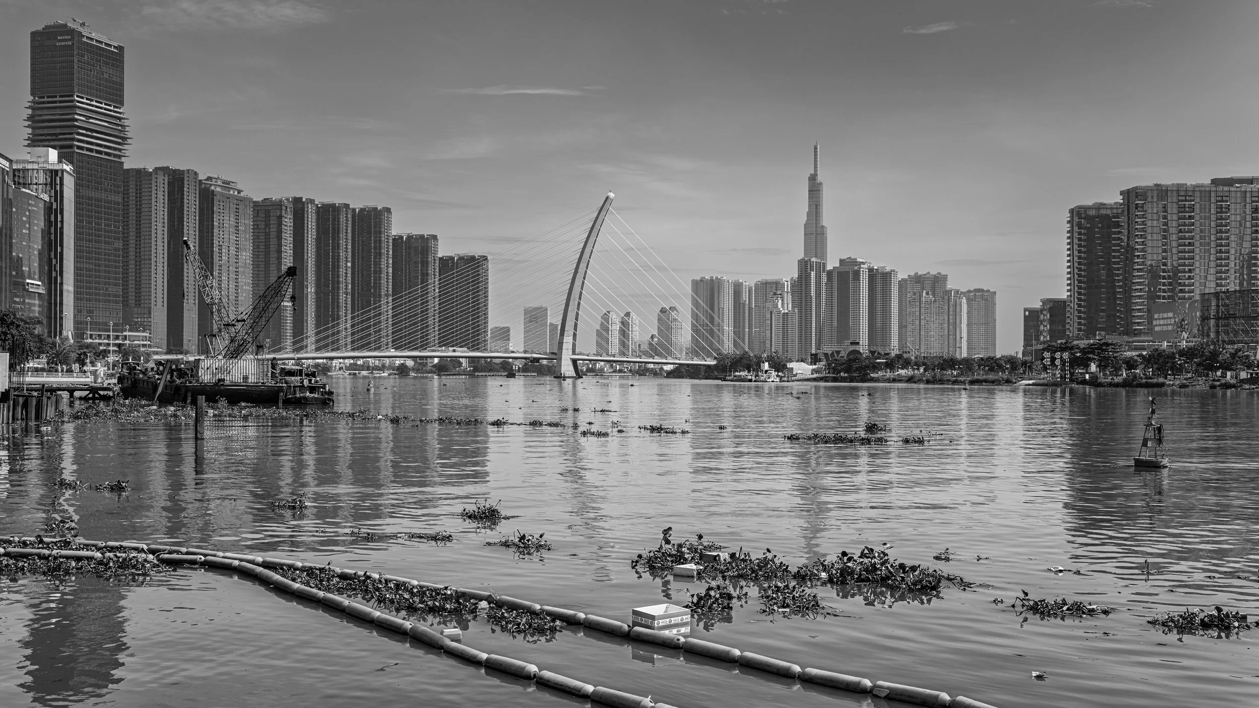 Black and white photo of a city skyline with tall buildings, a bridge over water, and a boat, with some floating debris in the water.