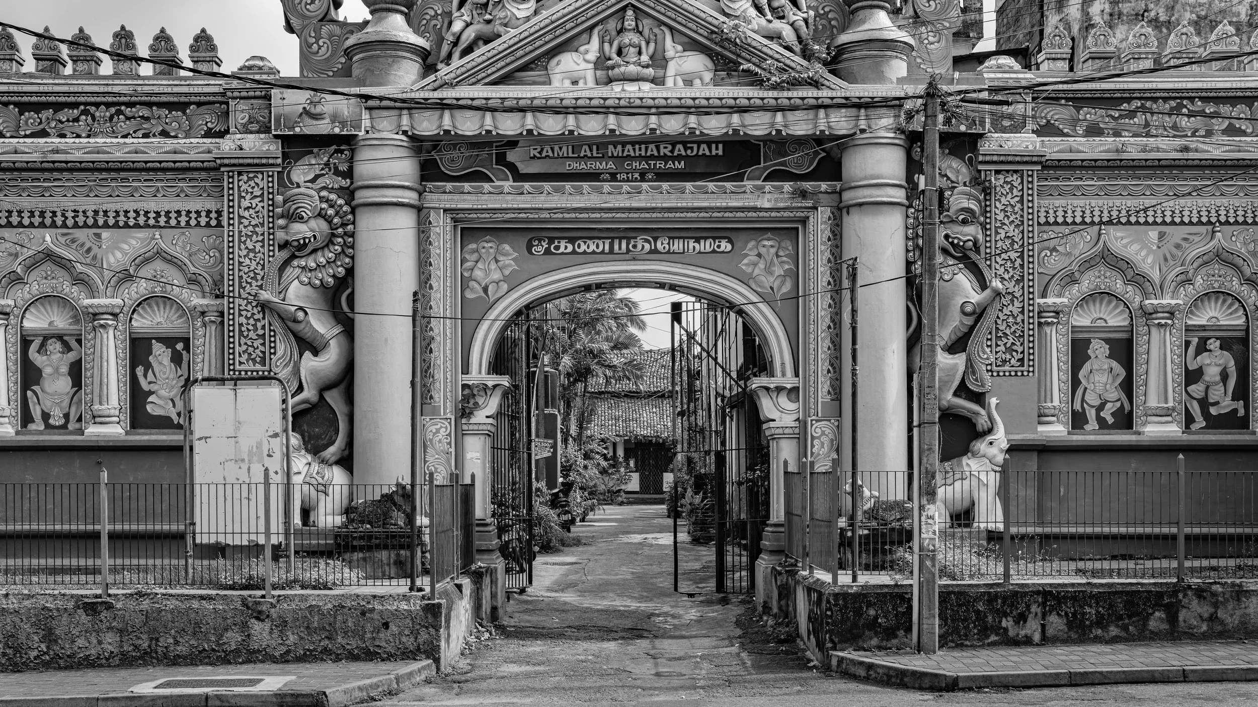 Black and white photo of an ornate temple entrance with intricate carvings and sculptures of mythological creatures and Hindu deities, including lions and elephants, with a gate leading into the temple grounds.