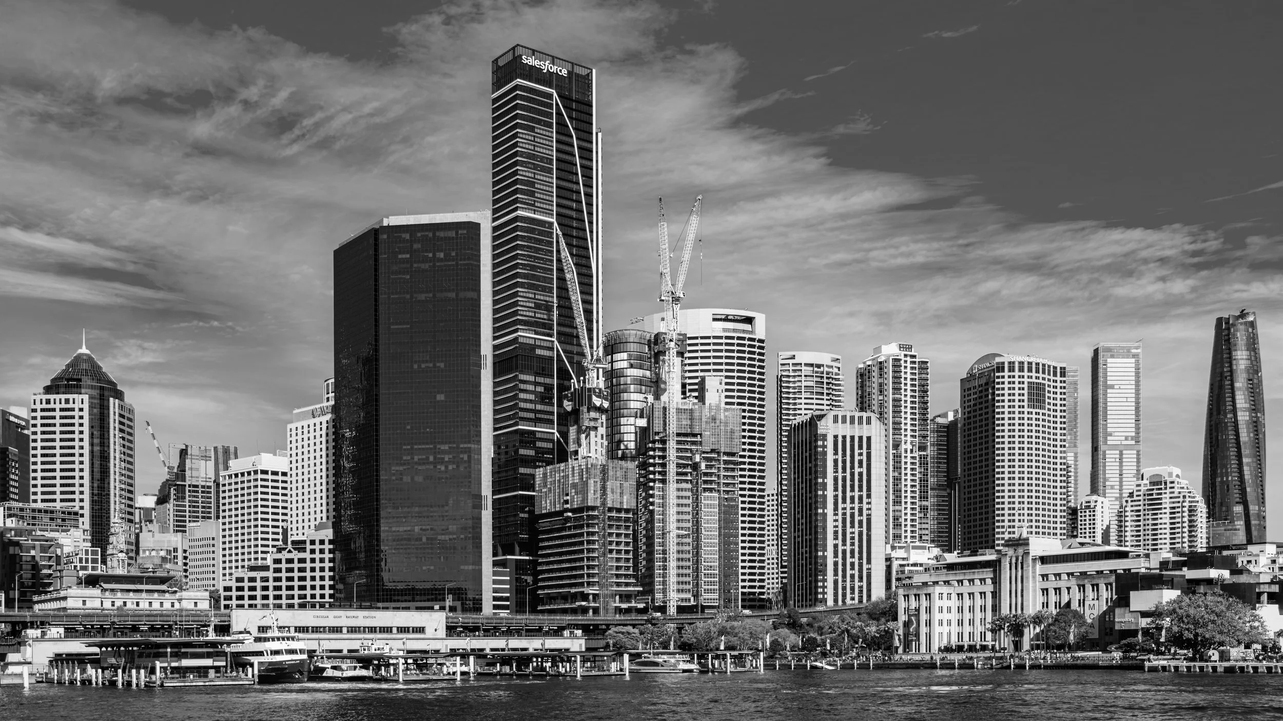 Black and white photograph of a city skyline with numerous tall skyscrapers, including the Salesforce Tower, under a partly cloudy sky, with water and boats in the foreground.