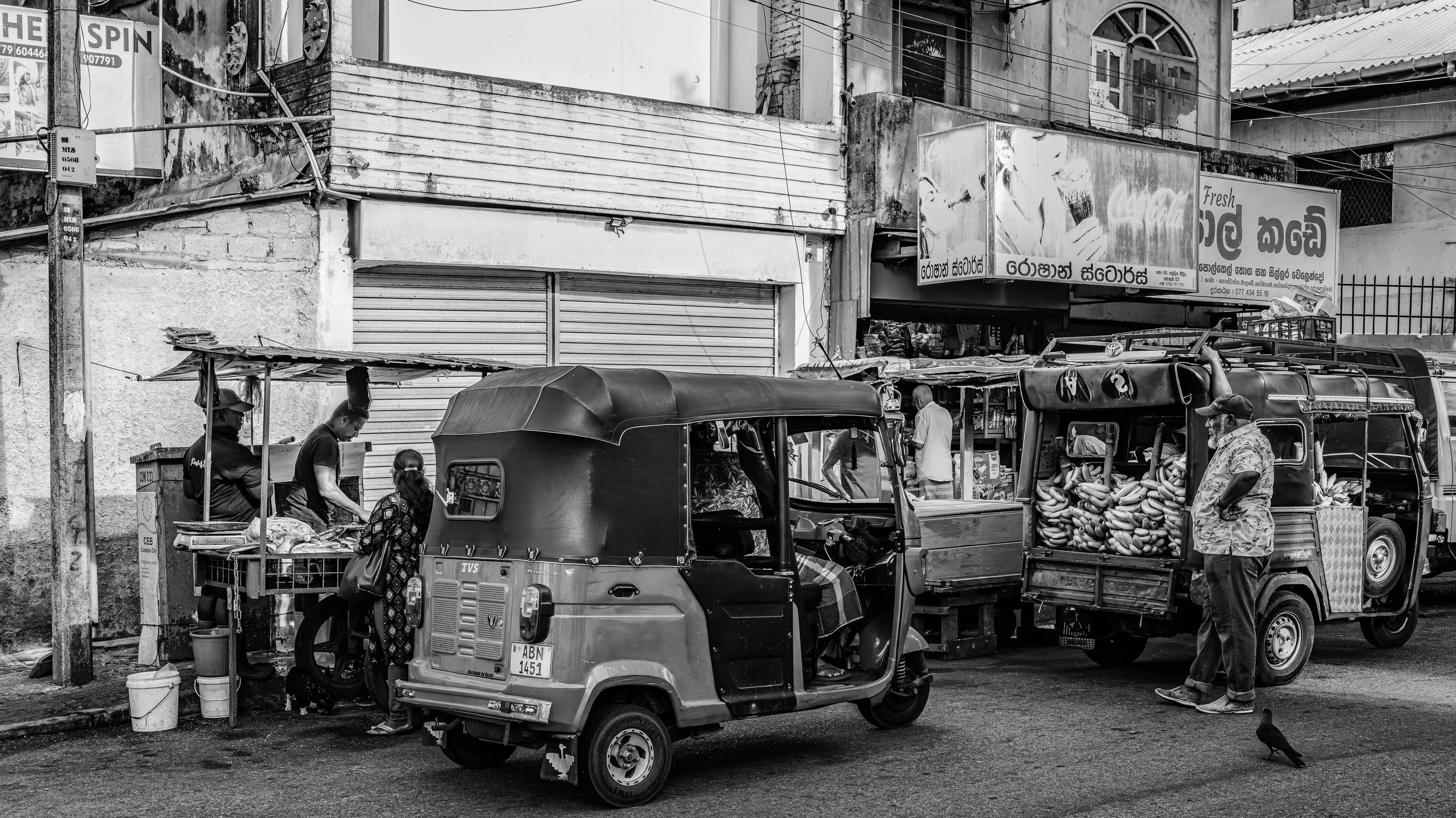 Street scene with three auto rickshaws and three people, two women and one man. One woman is wearing a patterned dress and carrying a bag. The man is wearing a floral shirt and a cap. The auto rickshaws are loaded with bananas and goods, and shop sig
