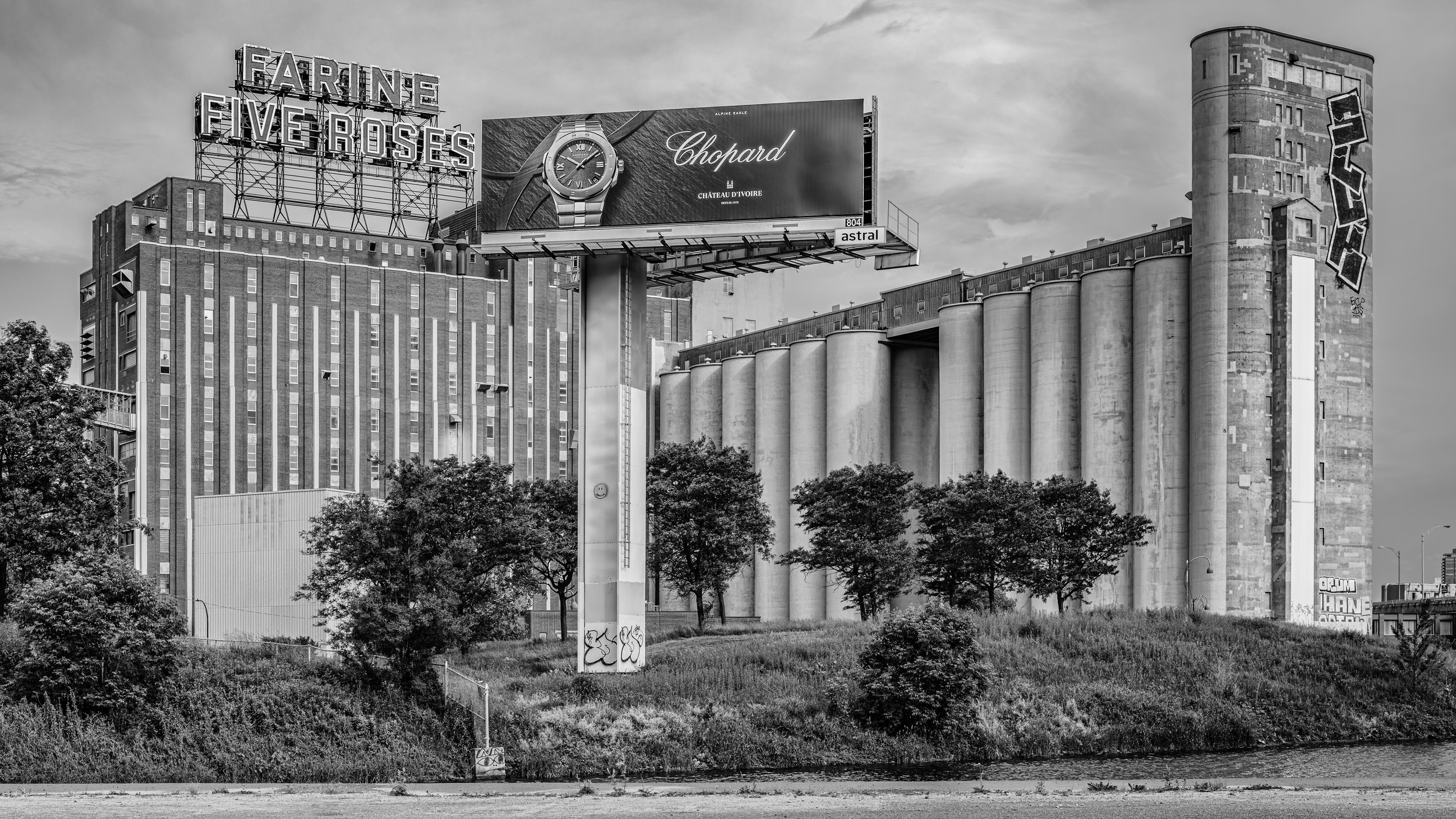 Black and white photo of city buildings with a billboard advertising Chopard watch, a large grain silo in the background, and trees in the foreground.
