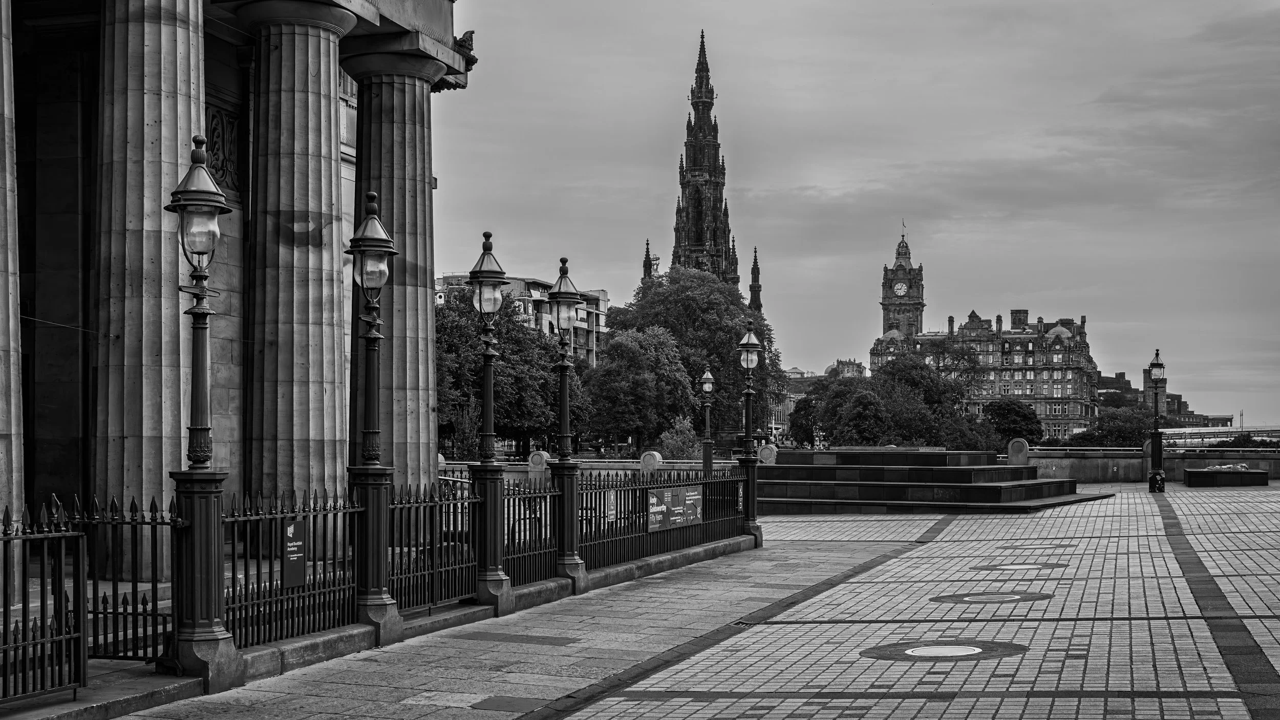 Black and white photo of a cityscape with historic buildings, trees, and street lamps, including a tall spire in the distance.
