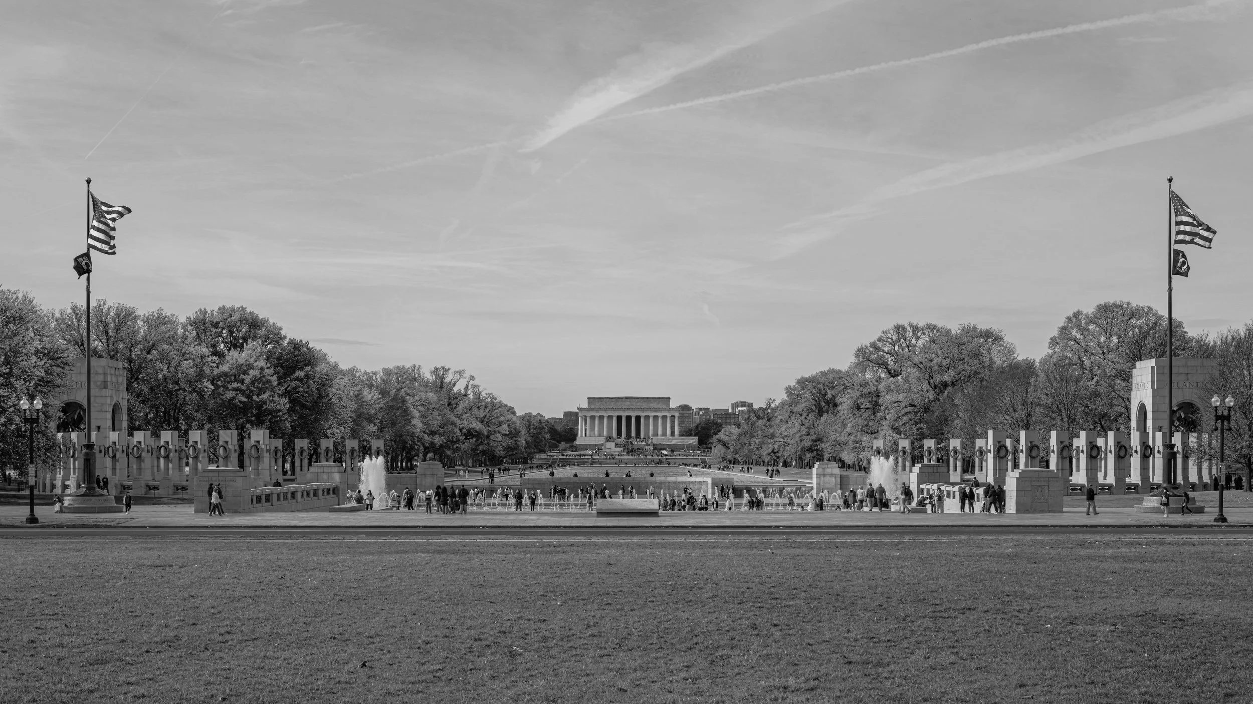 A black and white photograph of the National Mall in Washington D.C., featuring the Lincoln Memorial in the background, American flags on tall flagpoles, fountains, trees, and visitors walking around.