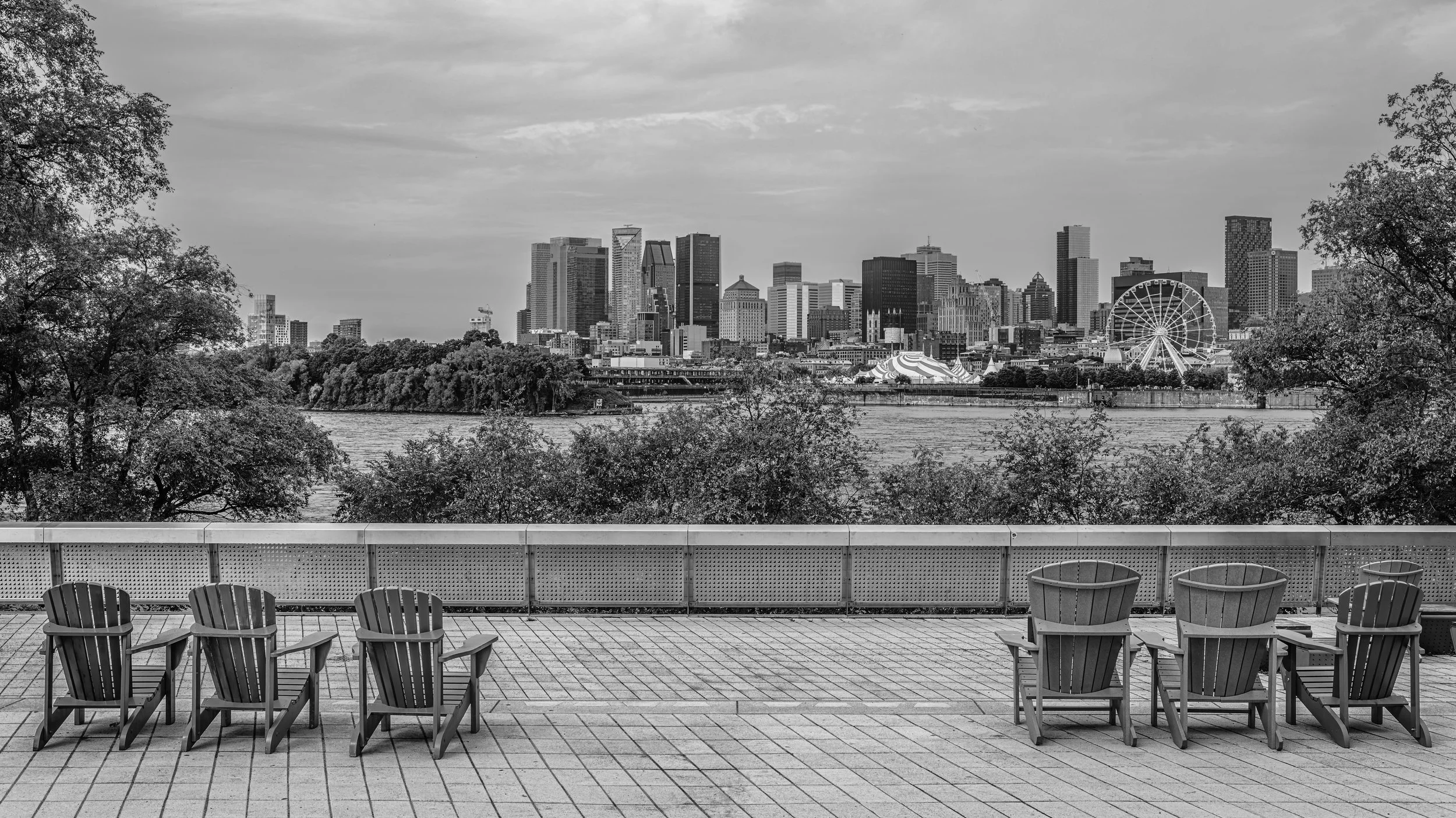 View of a city skyline with tall buildings and a Ferris wheel in the distance, seen from a waterfront area with chairs and trees in the foreground.
