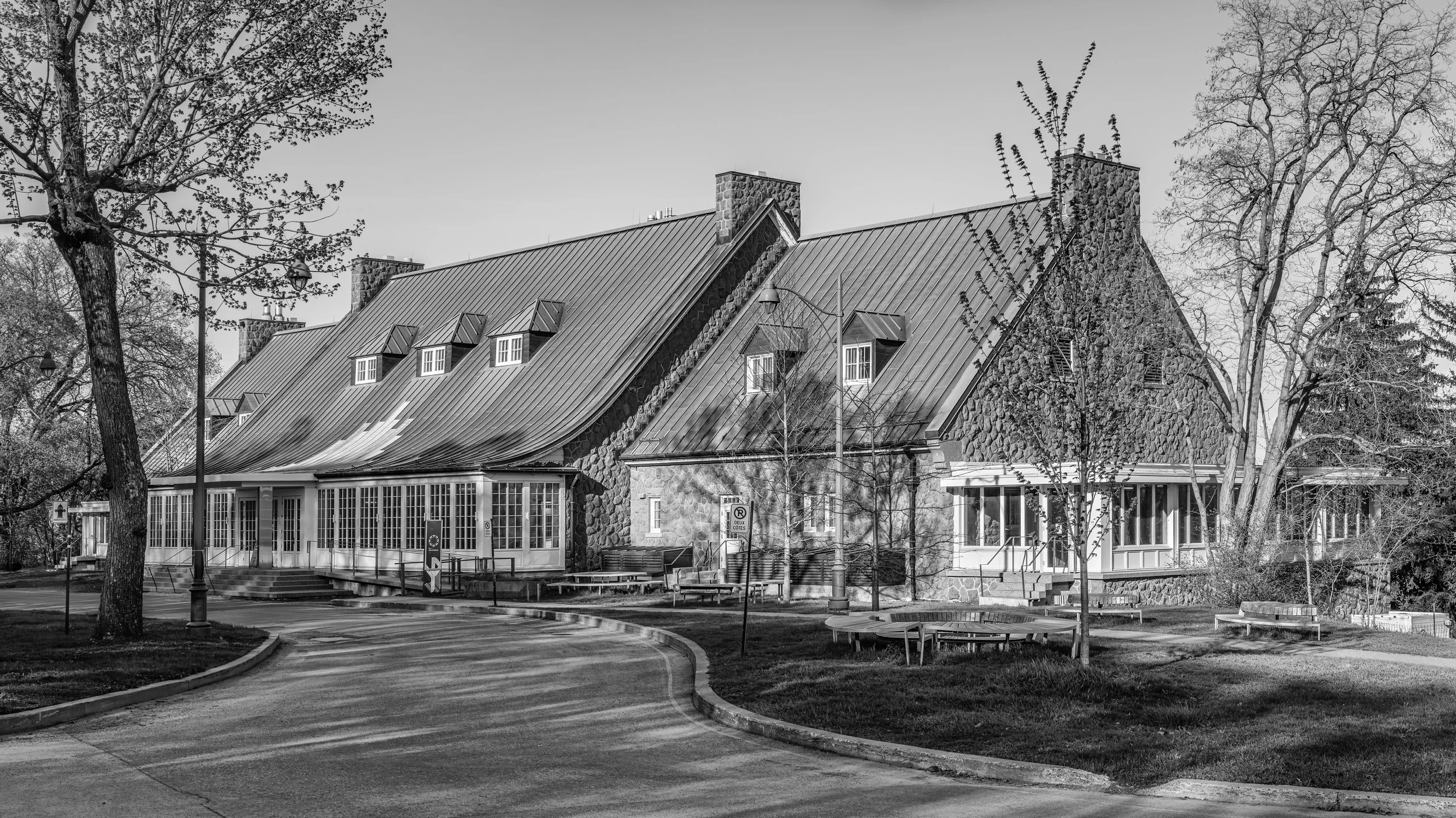 Black and white photo of a building with a steep metal roof, stone walls, and small dormer windows, surrounded by trees and a park-like setting.