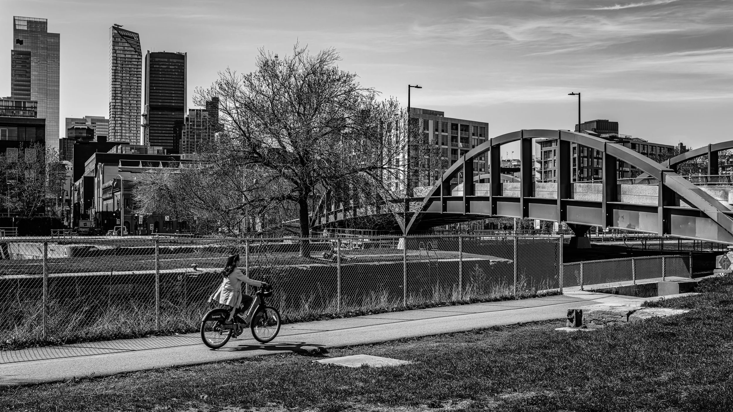 A black and white photo of a cityscape with tall buildings in the background and a park in the foreground. A pedestrian bridge crosses over a fenced area, and a child is riding a bicycle on a paved path in the park.