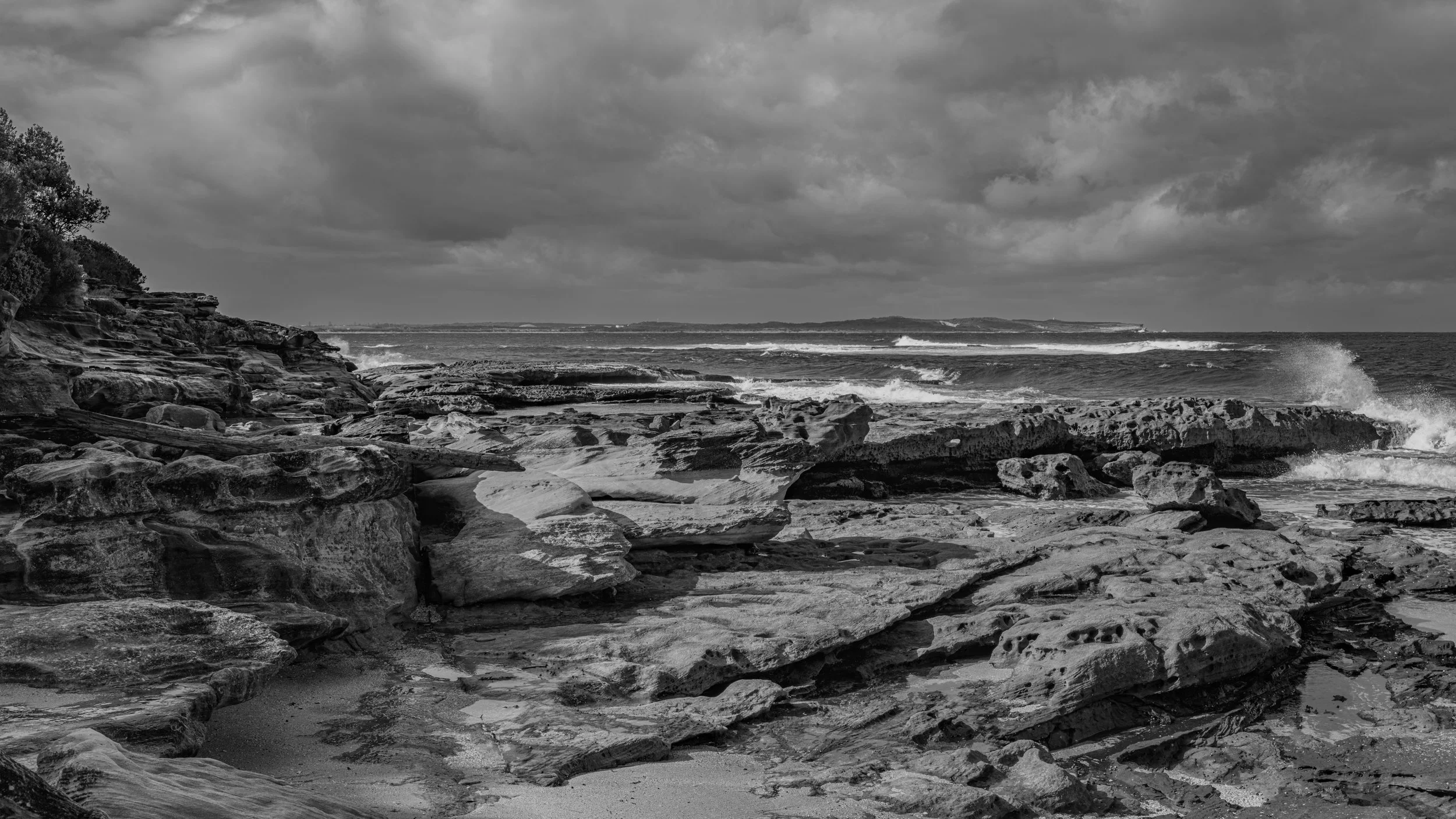 Coastal scene with rocky shoreline, ocean waves, and cloudy sky in black and white.