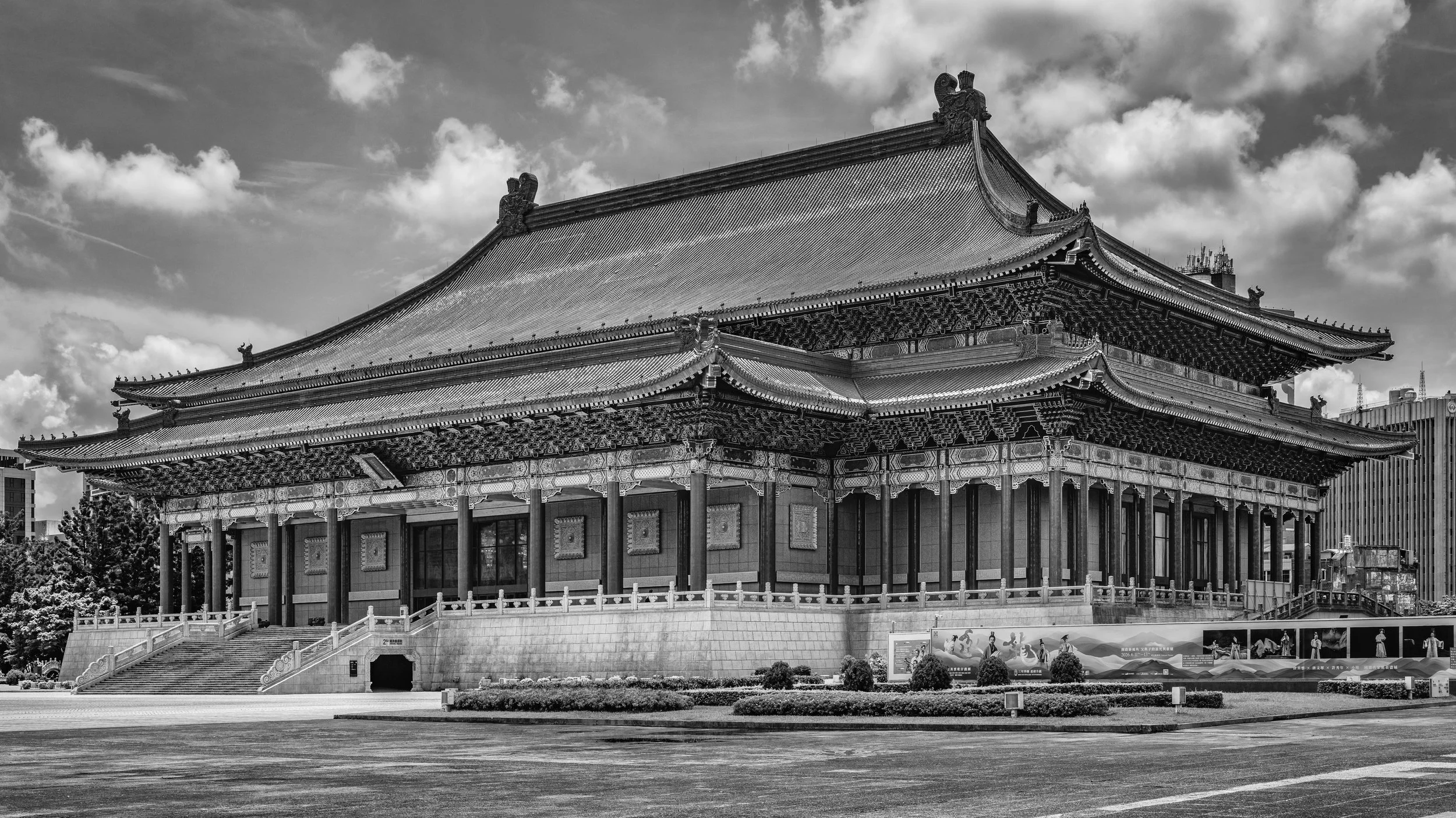 Black and white photo of a large traditional Asian temple with ornate roof decorations, located in an urban area with modern buildings nearby, under a partly cloudy sky.