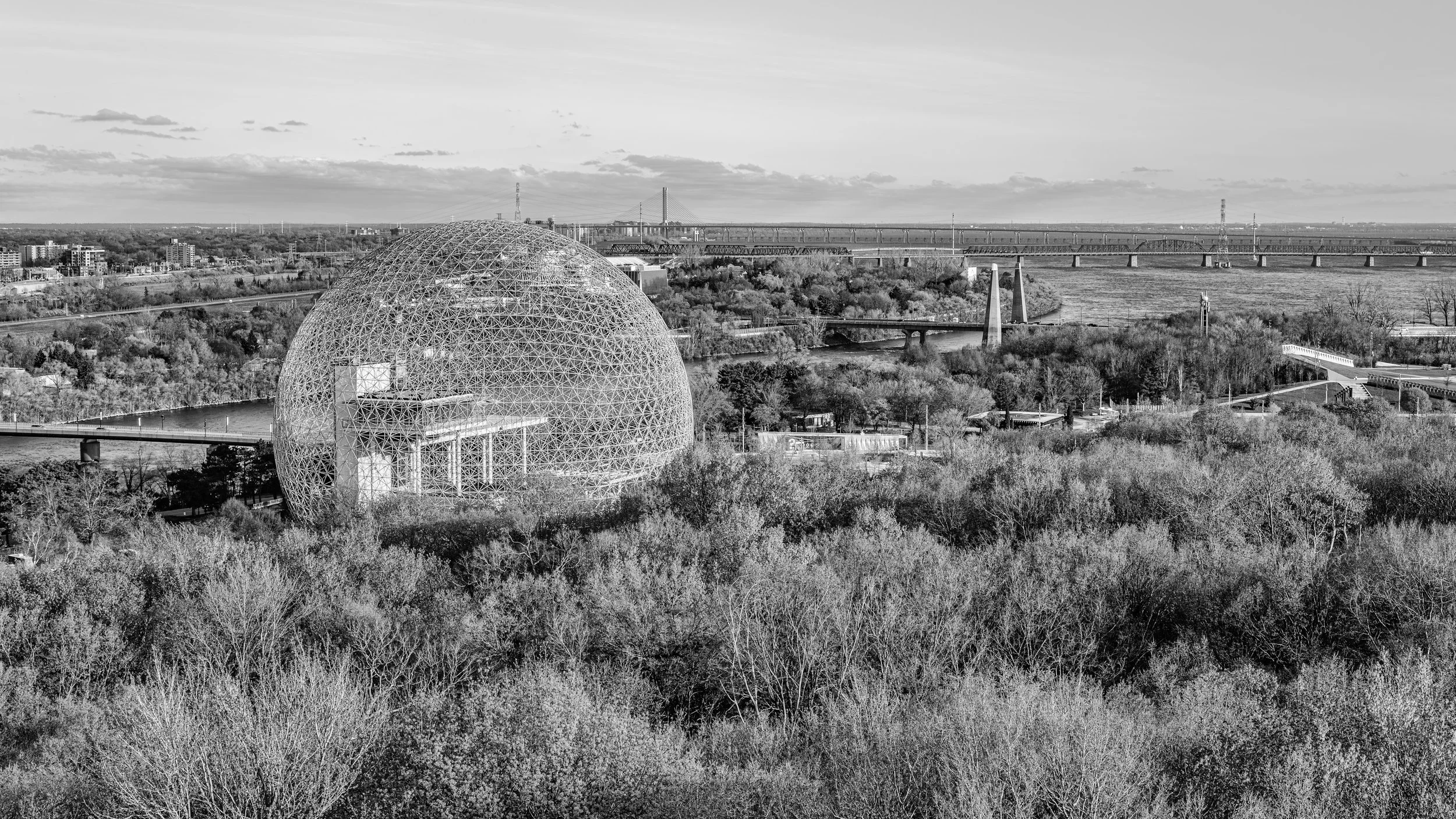 Black and white landscape featuring a large geodesic dome structure in the foreground, surrounded by trees, with bridges and a river in the background under a cloudy sky.