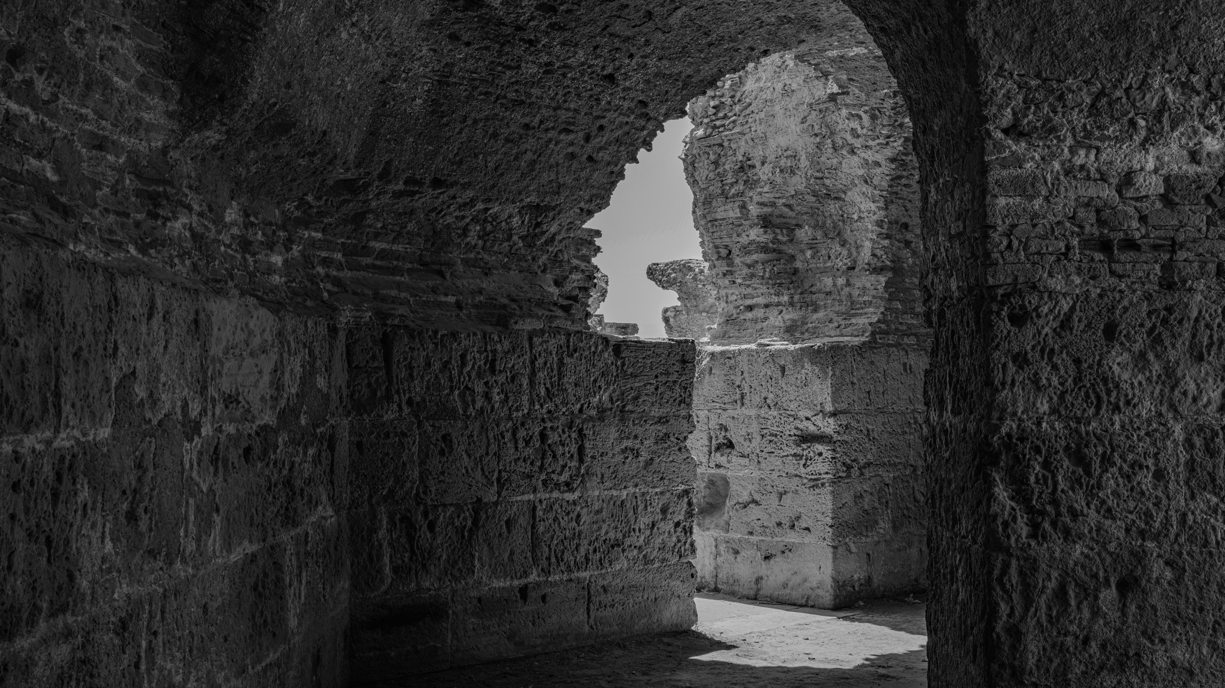 View of ancient stone ruins through an arched opening, with weathered stone walls and a distant view of sky and stone formations.