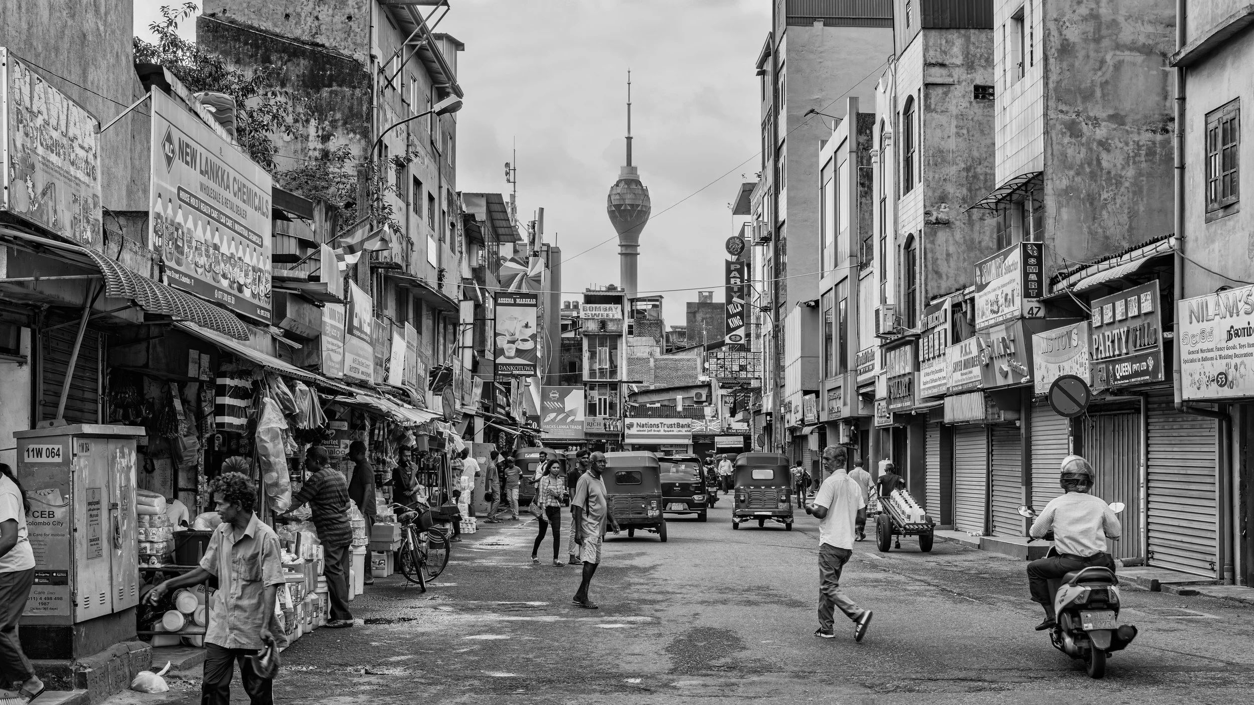 Street scene in Sri Lanka with people walking, parked tuk-tuks, and buildings with signs in local language. The Sky Tower is visible in the distance.