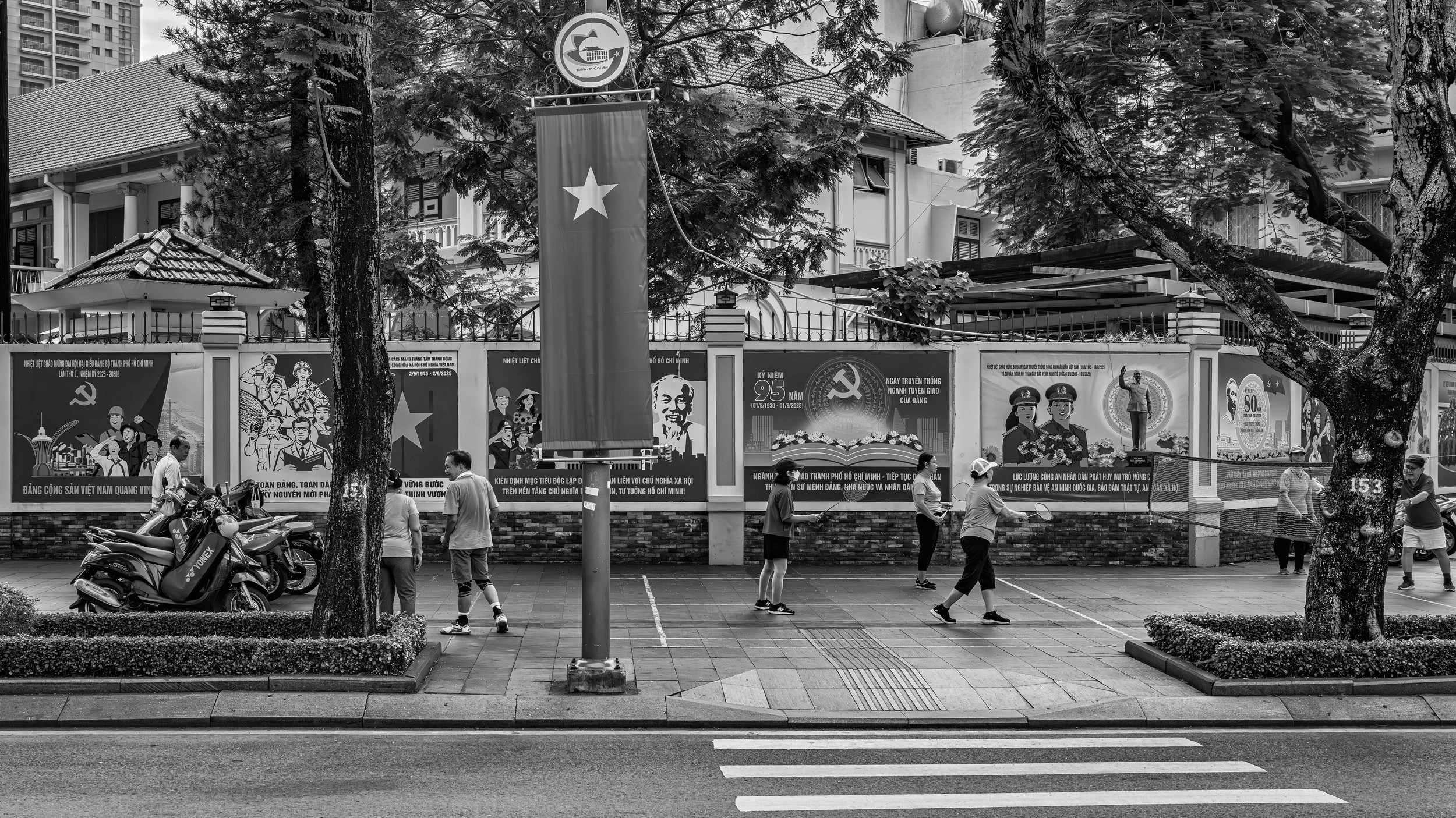 Black and white photo of people standing in front of a wall decorated with posters, a large flag with a star, and the communist hammer and sickle symbol. The scene is outdoors with trees and buildings visible in the background.
