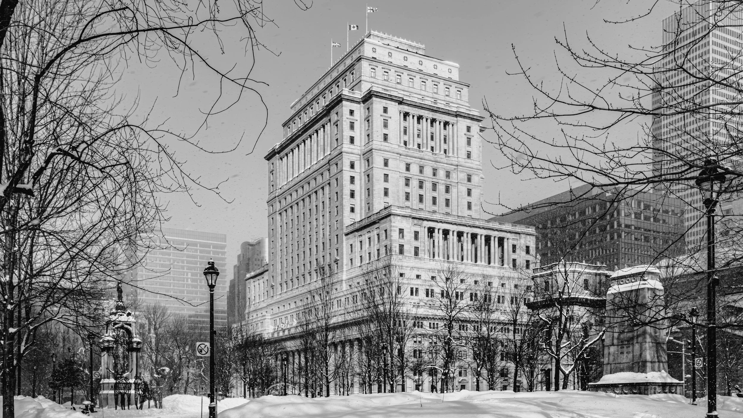 Black and white photograph of tall, historic city building with flag atop, surrounded by leafless trees and modern skyscrapers in the background, with snow on the ground and city street lamps visible.