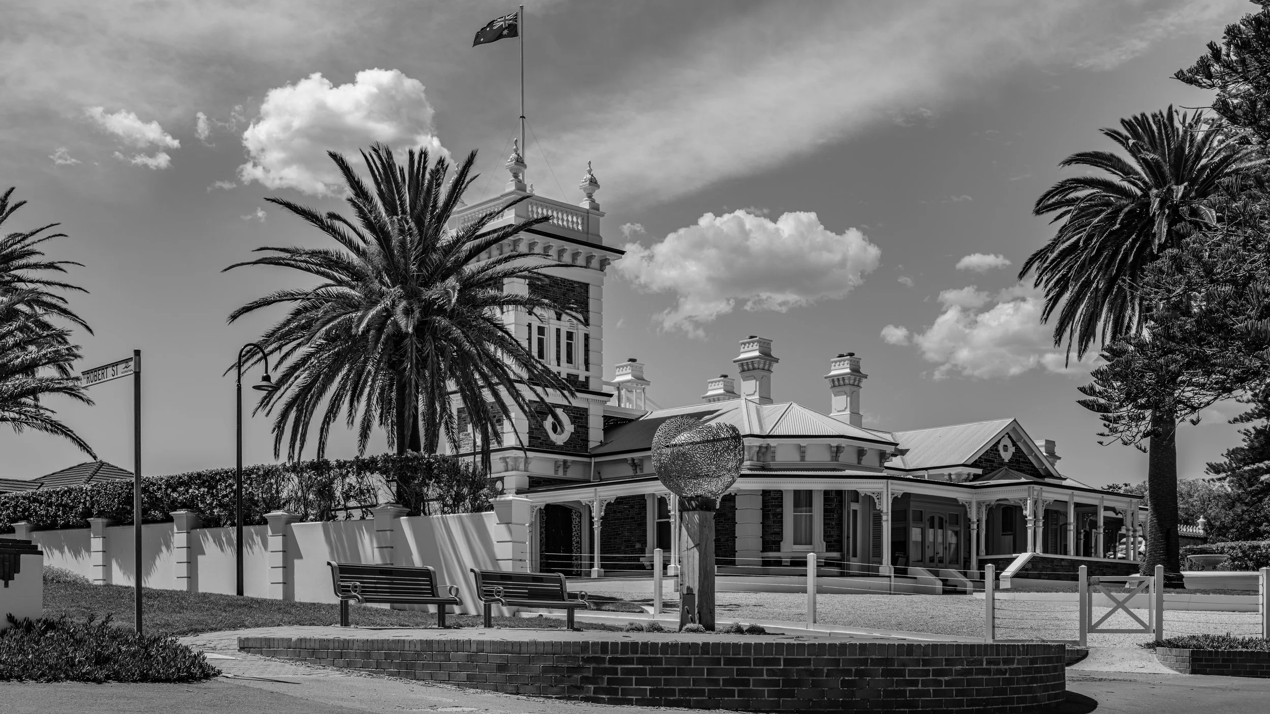 A historic building with Victorian architecture, featuring a prominent tower with an Australian flag on top, surrounded by palm trees, a park bench, and a brick and white fence. The photo is in black and white.