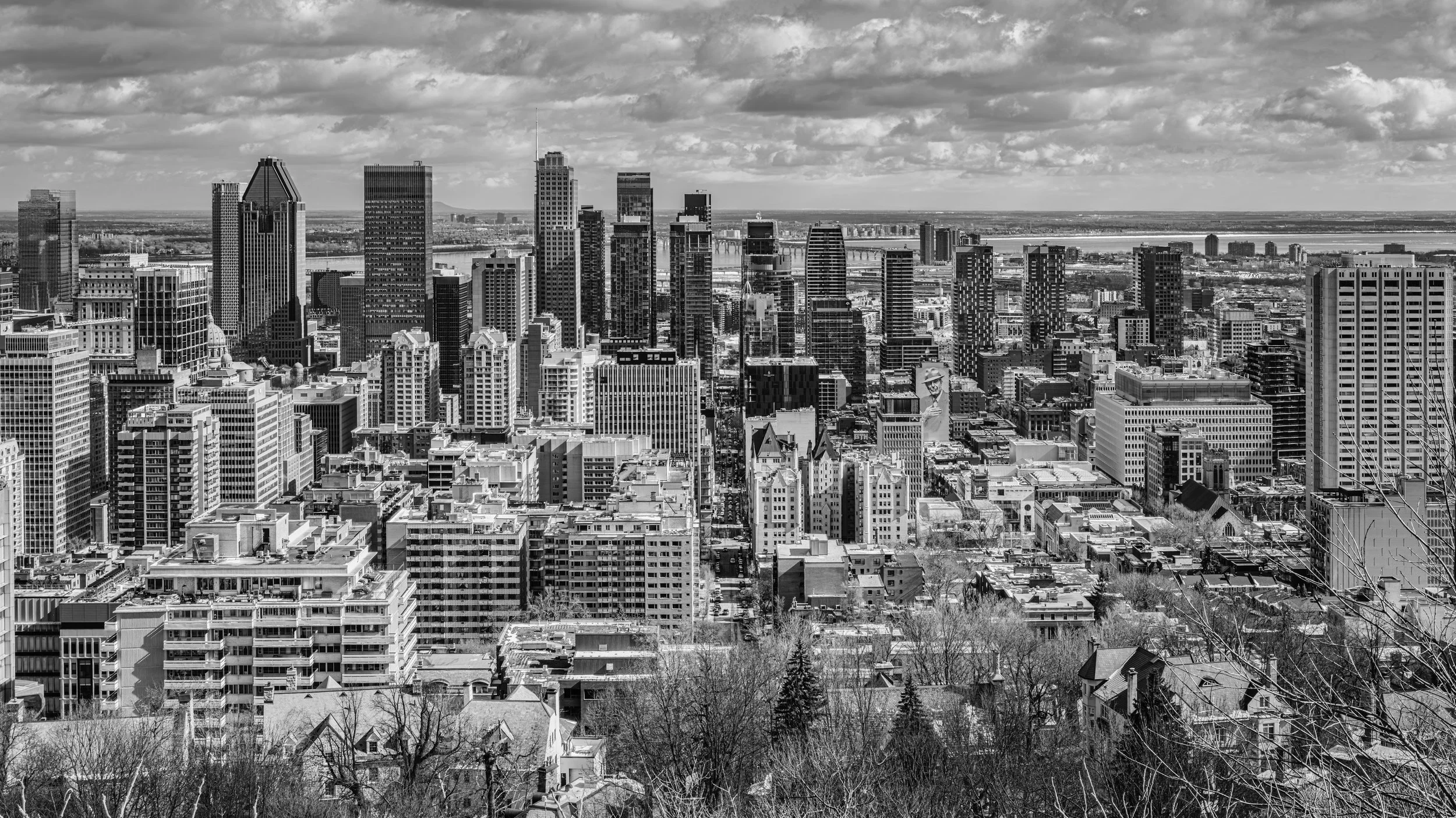 Black and white aerial view of a city skyline with tall office buildings, residential apartments, and a river in the background under a cloudy sky.