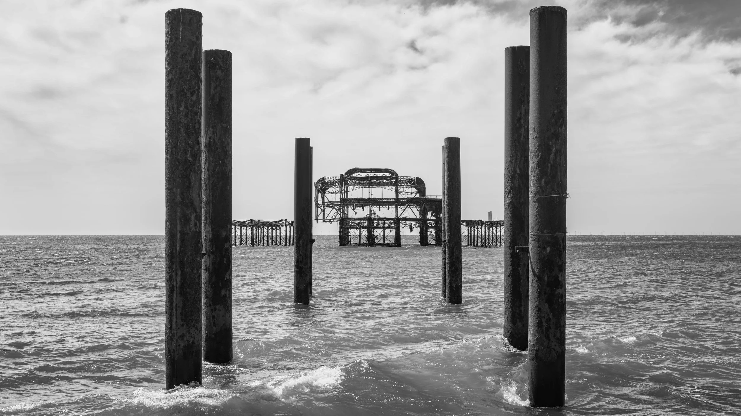Black and white photo of a beach with several old, weathered poles in the water and a partially collapsed pier structure in the background.