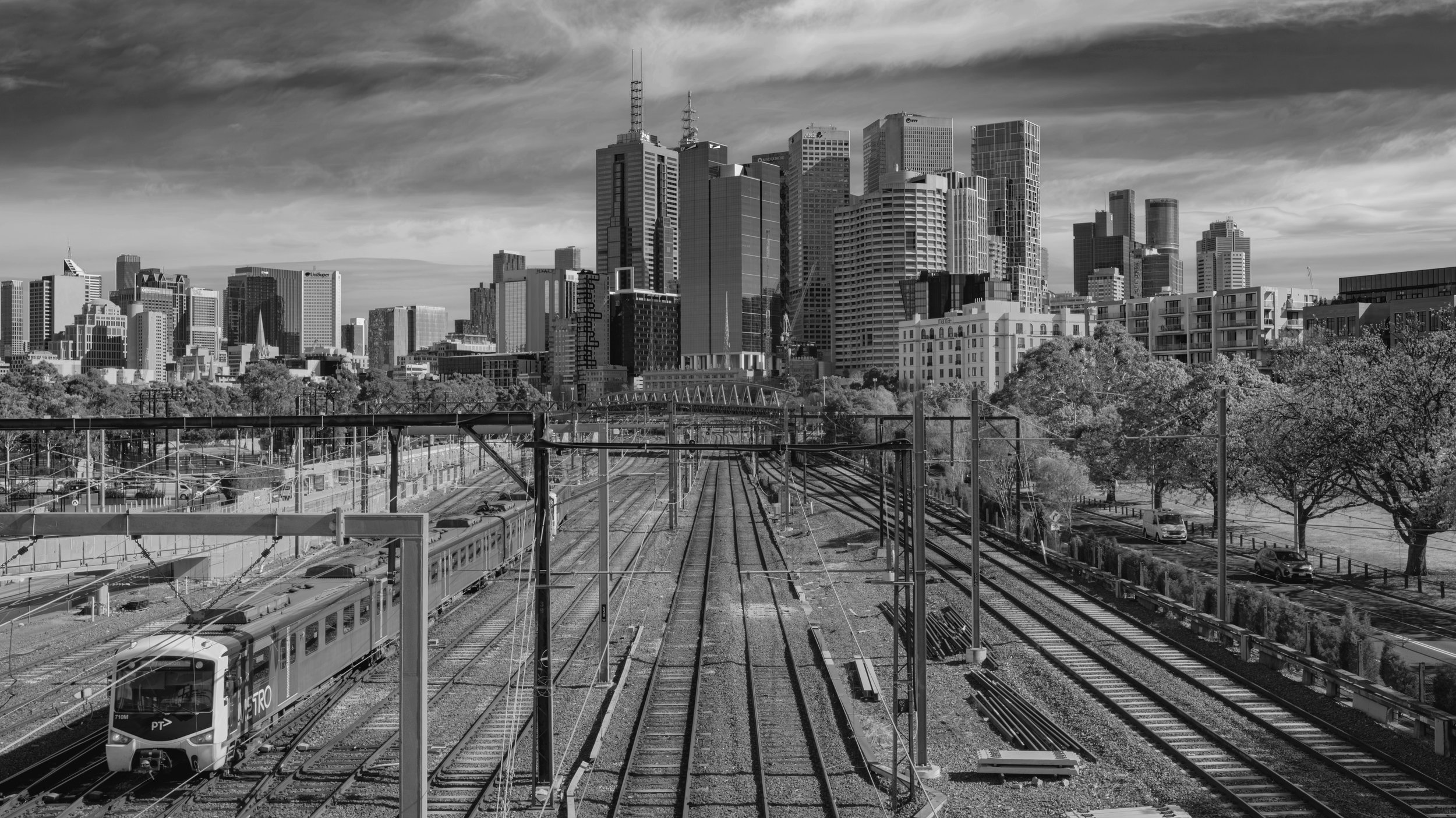 Black and white photo of a city skyline with tall buildings, railway tracks in the foreground, and a train moving on the tracks. Trees are visible along the right side of the tracks.