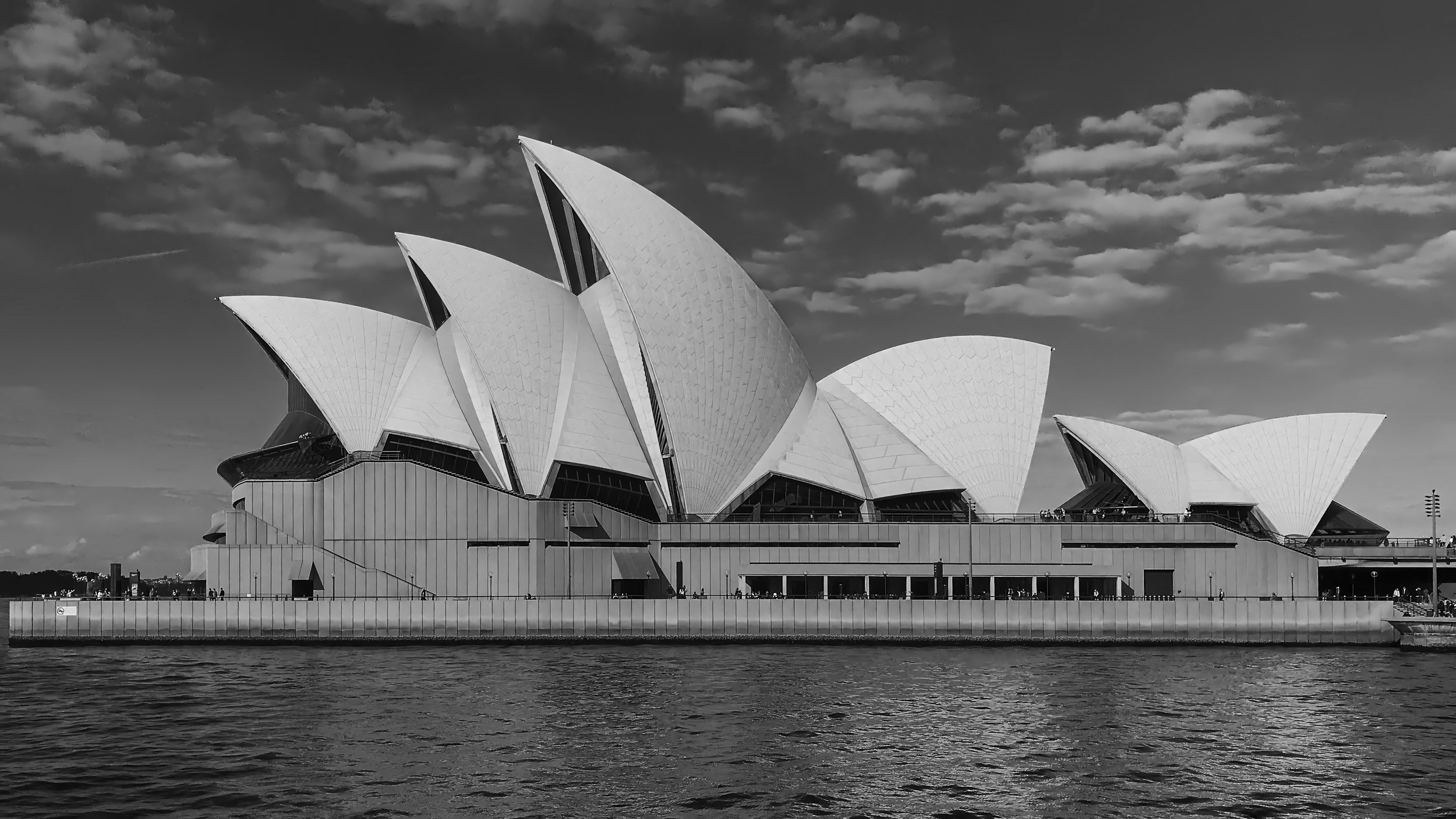 A black and white photograph of the Sydney Opera House with its distinctive sail-like roof structure, situated along the harbor with water in the foreground and cloudy sky above.