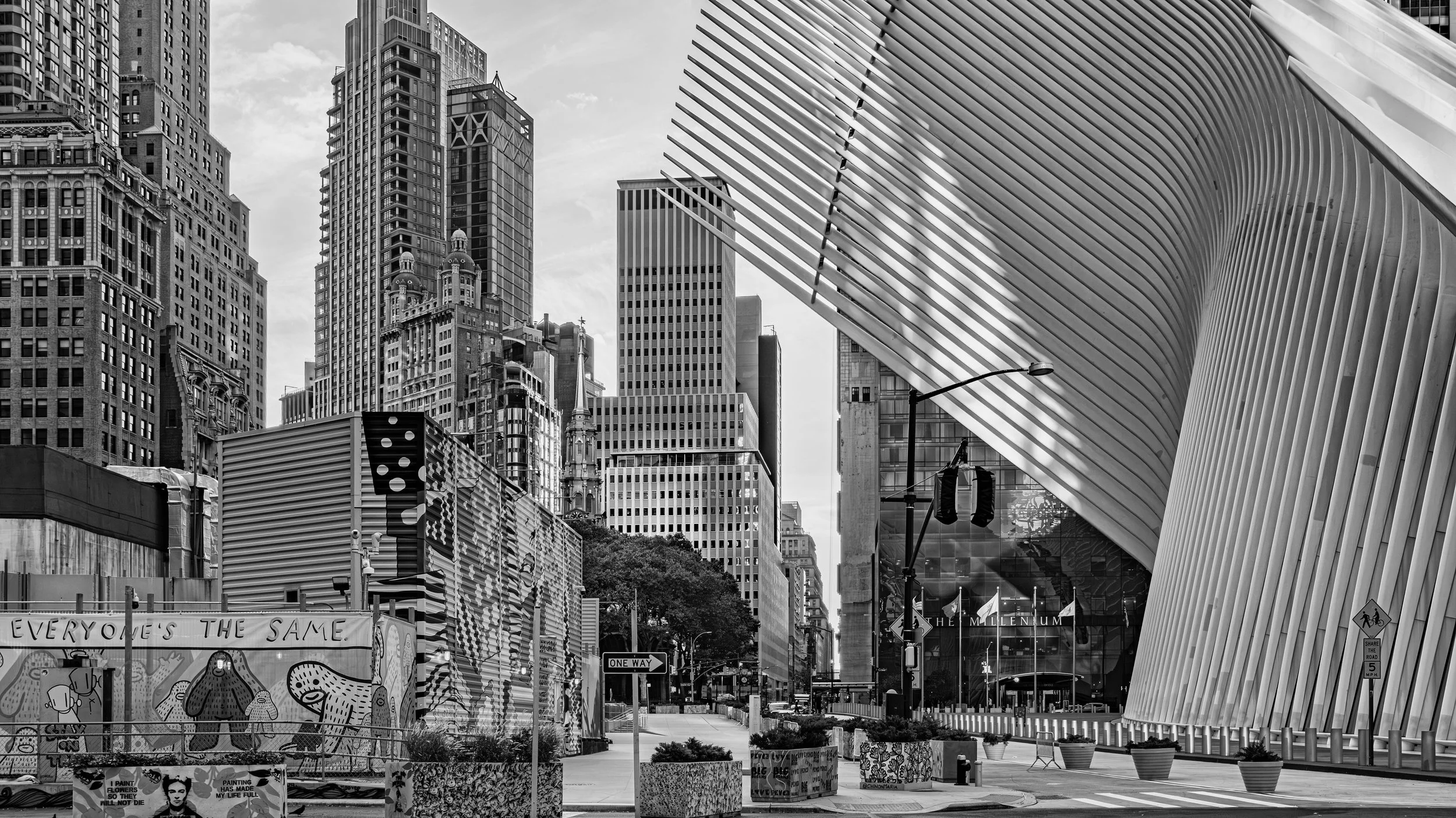 Black and white photo of a city street with tall skyscrapers and modern architectural buildings, including a distinctive curved, striped building on the right. Street signs and potted plants are visible in the foreground. Manhattan, New York, USA.