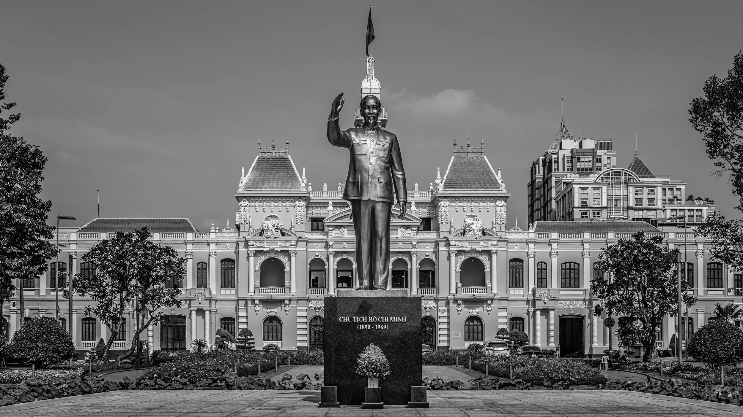 Statue of Ho Chi Minh in front of a colonial-style building with a garden in the foreground, trees, and a partly cloudy sky.