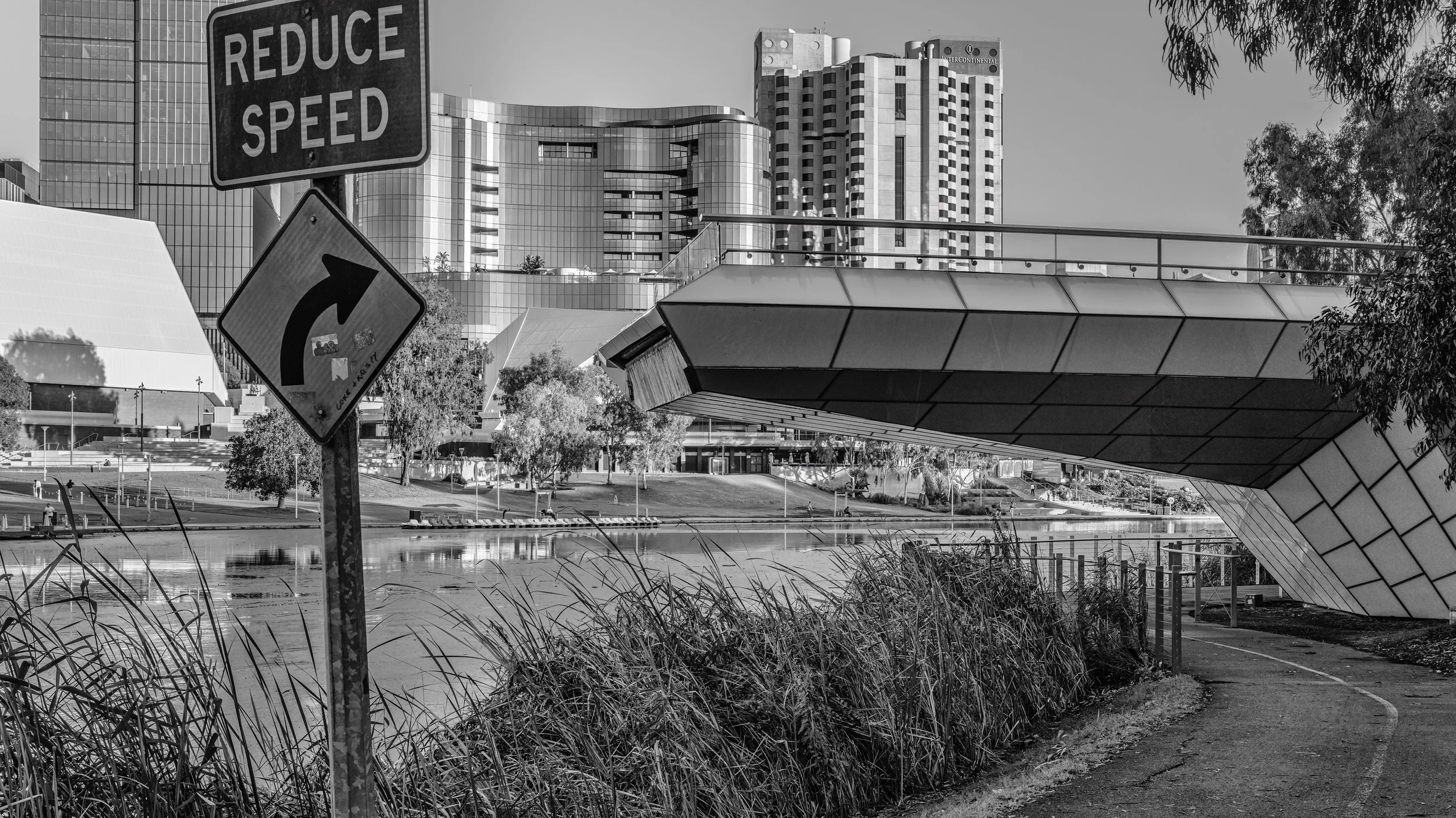 Black and white photo of an urban park with modern buildings in the background. There are road signs, including one that says 'Reduce Speed' and a curved arrow indicating a turn. Adelaide, Australia.
