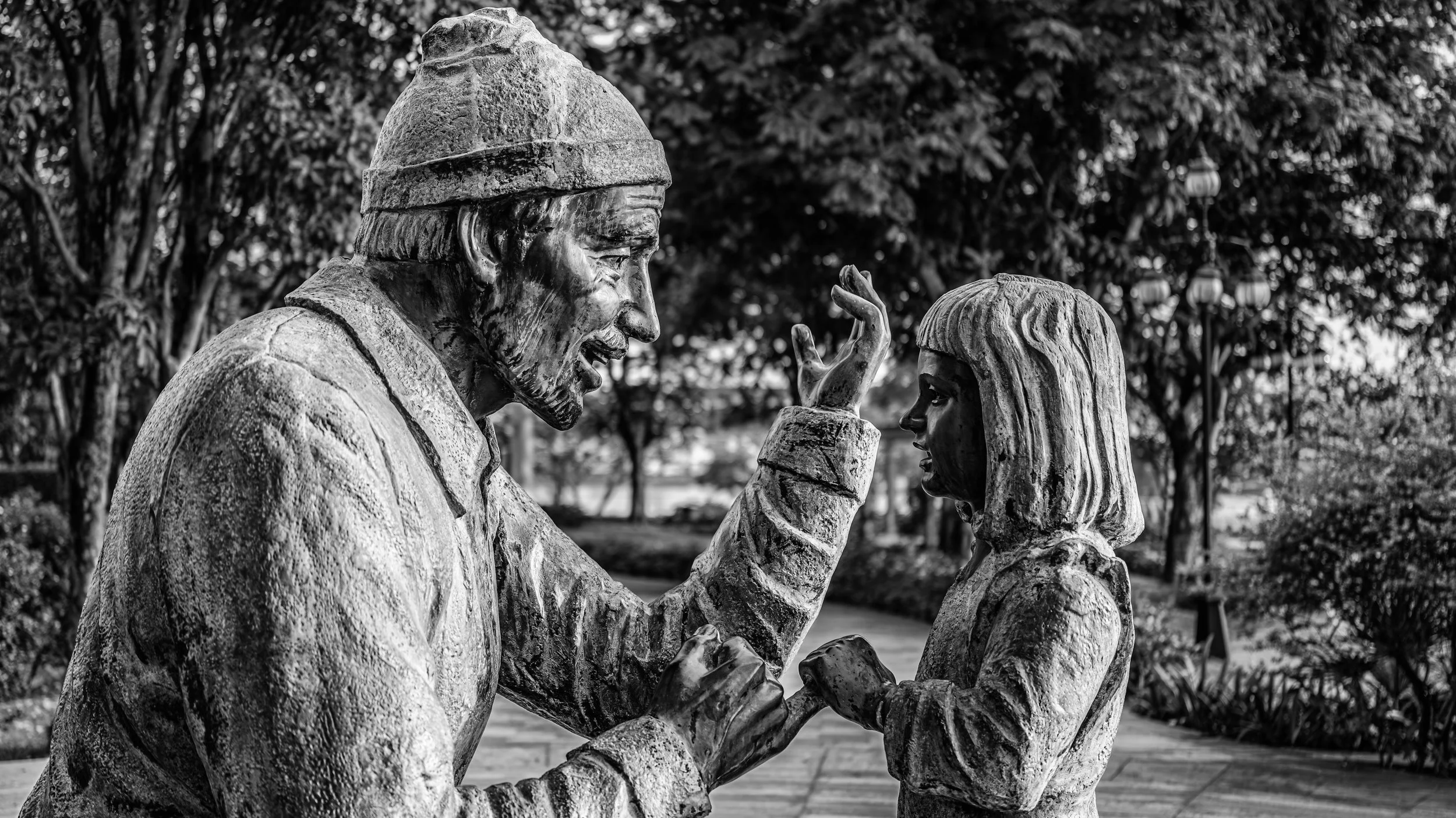 Black and white photo of a sculpture of an older man and a young girl. They are facing each other with their hands touching. The scene is outdoors with trees and lanterns in the background.