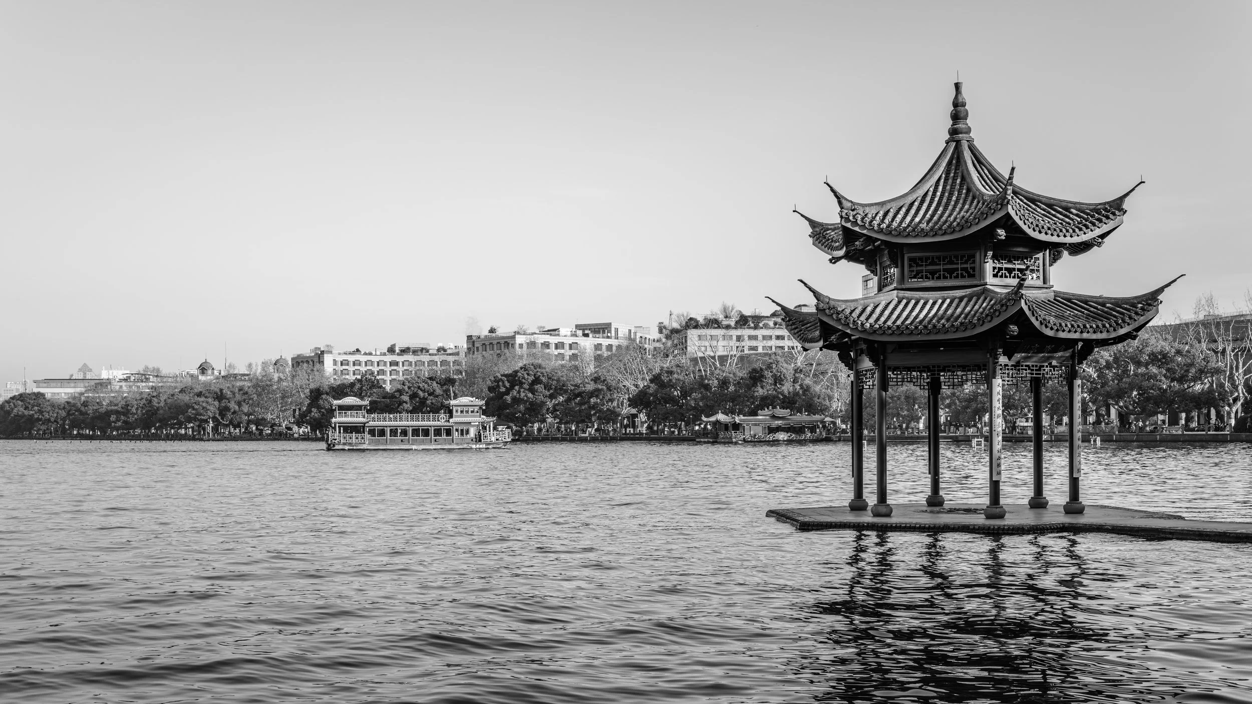 A traditional Chinese pavilion with ornate curved roof and pillars, situated on the edge of a body of water, with a cityscape and trees in the background, in black and white.