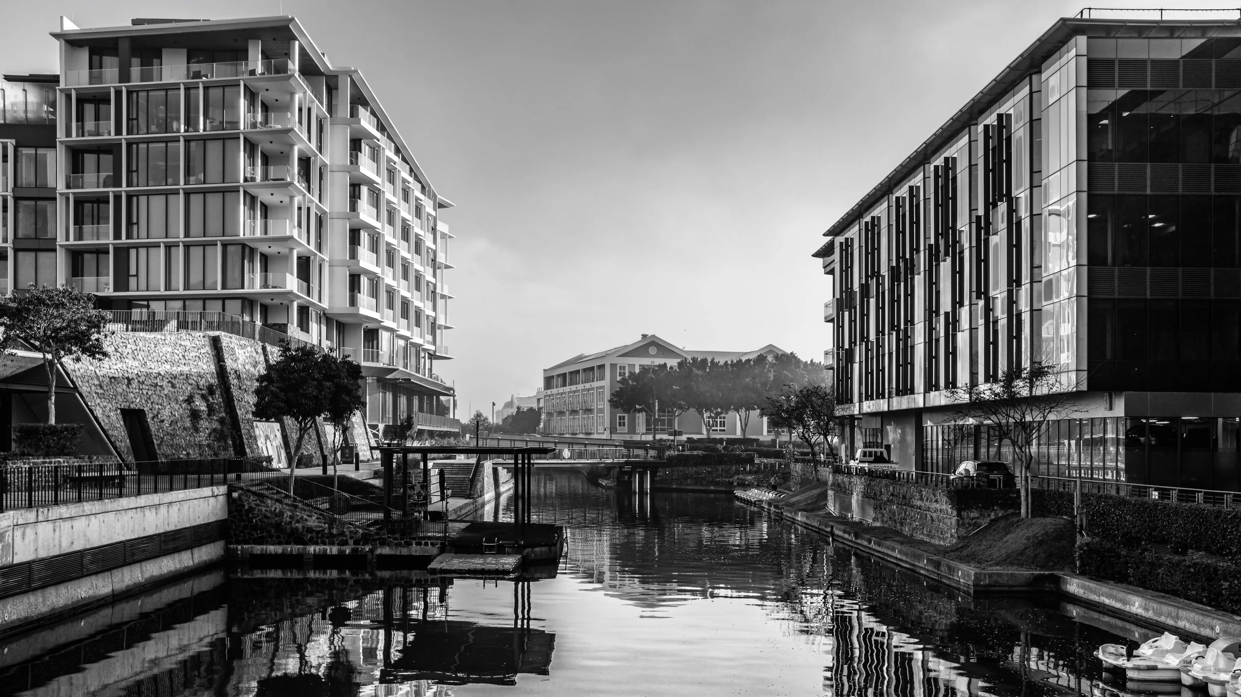 Black and white photo of modern apartment buildings overlooking a canal with small boats, trees, and a bridge in an urban setting. Cape Town, South Africa.