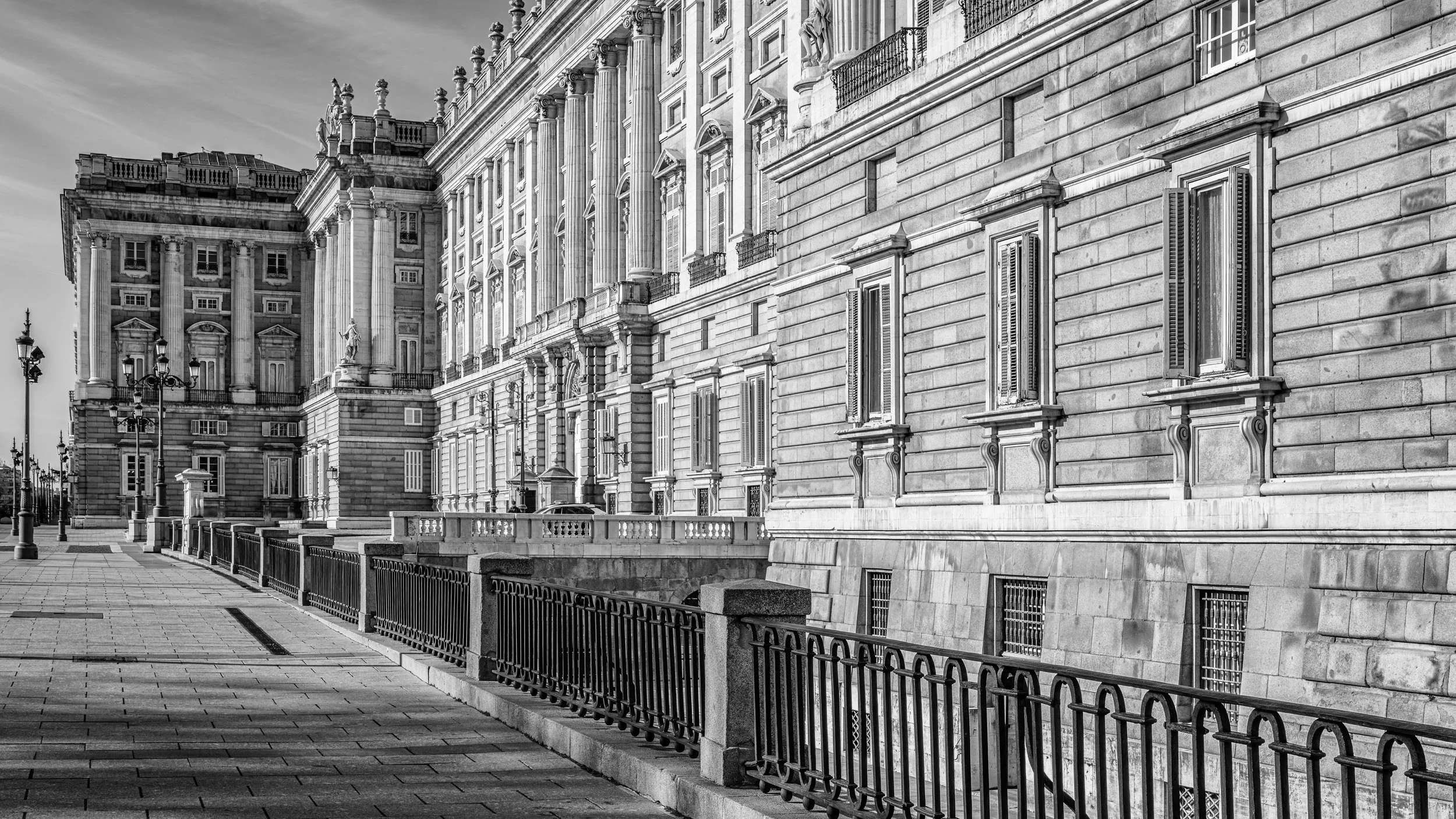 Black and white photograph of a historic building with ornate architectural details, multiple windows with shutters, and decorative columns, along a paved walkway with street lamps and a metal fence.