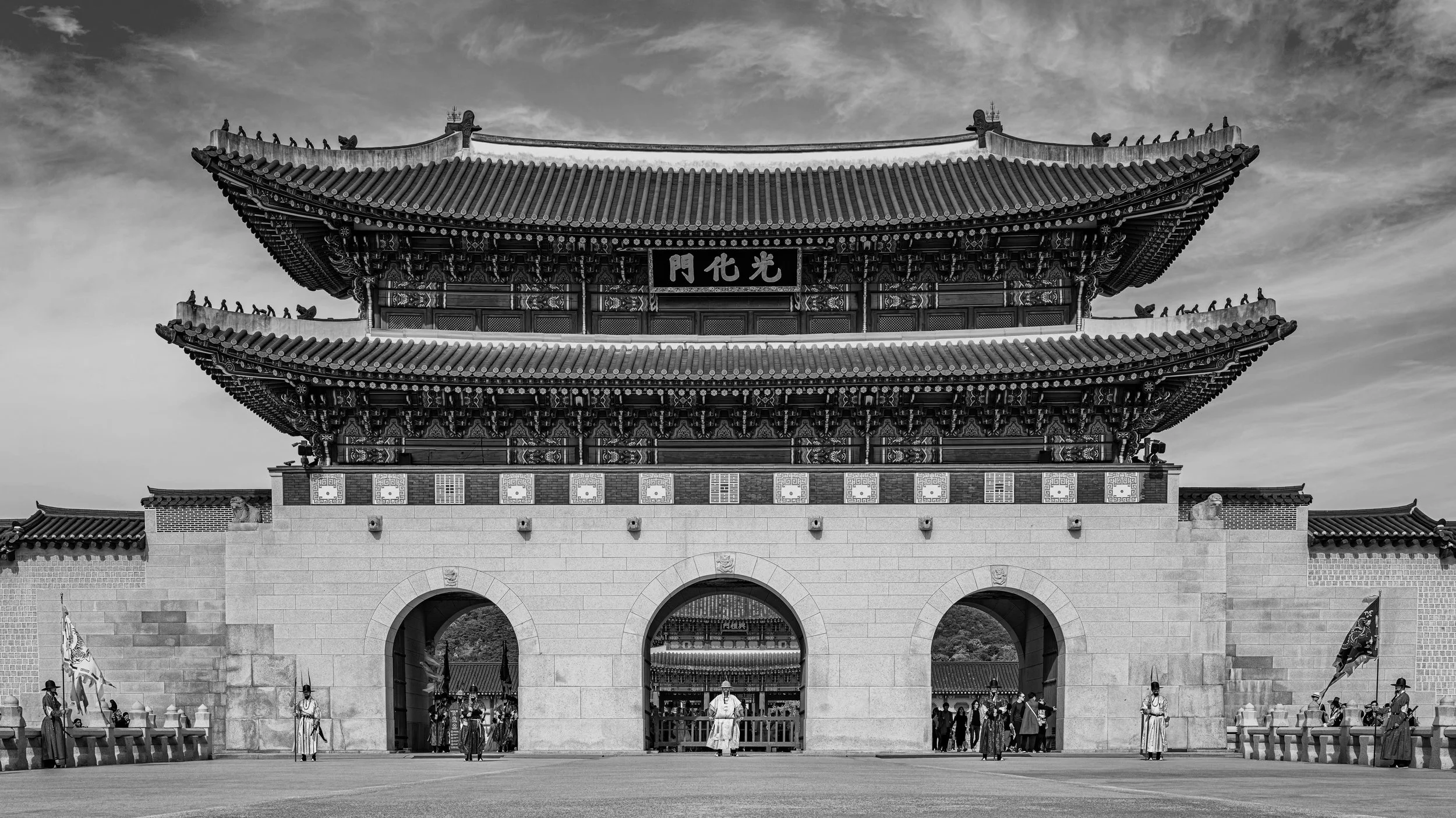 Black and white photo of a traditional Chinese gate with ornate roof, decorated with intricate patterns and a sign with Chinese characters, with people in traditional attire and soldiers standing guard in front.