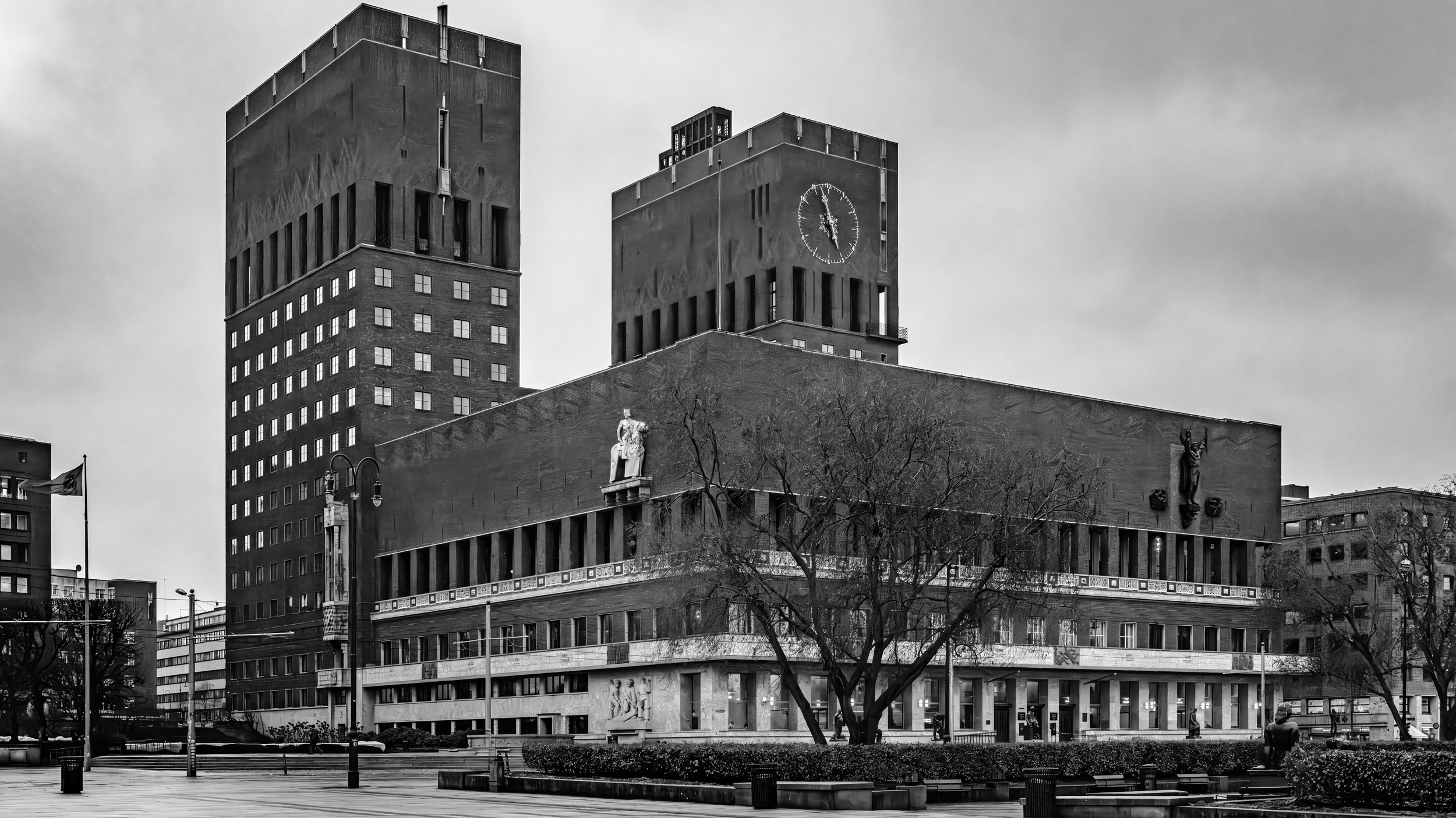 Black and white photo of a modern building with two towers, one with a clock, and statues on the façade, with bare trees in front and cloudy sky overhead.