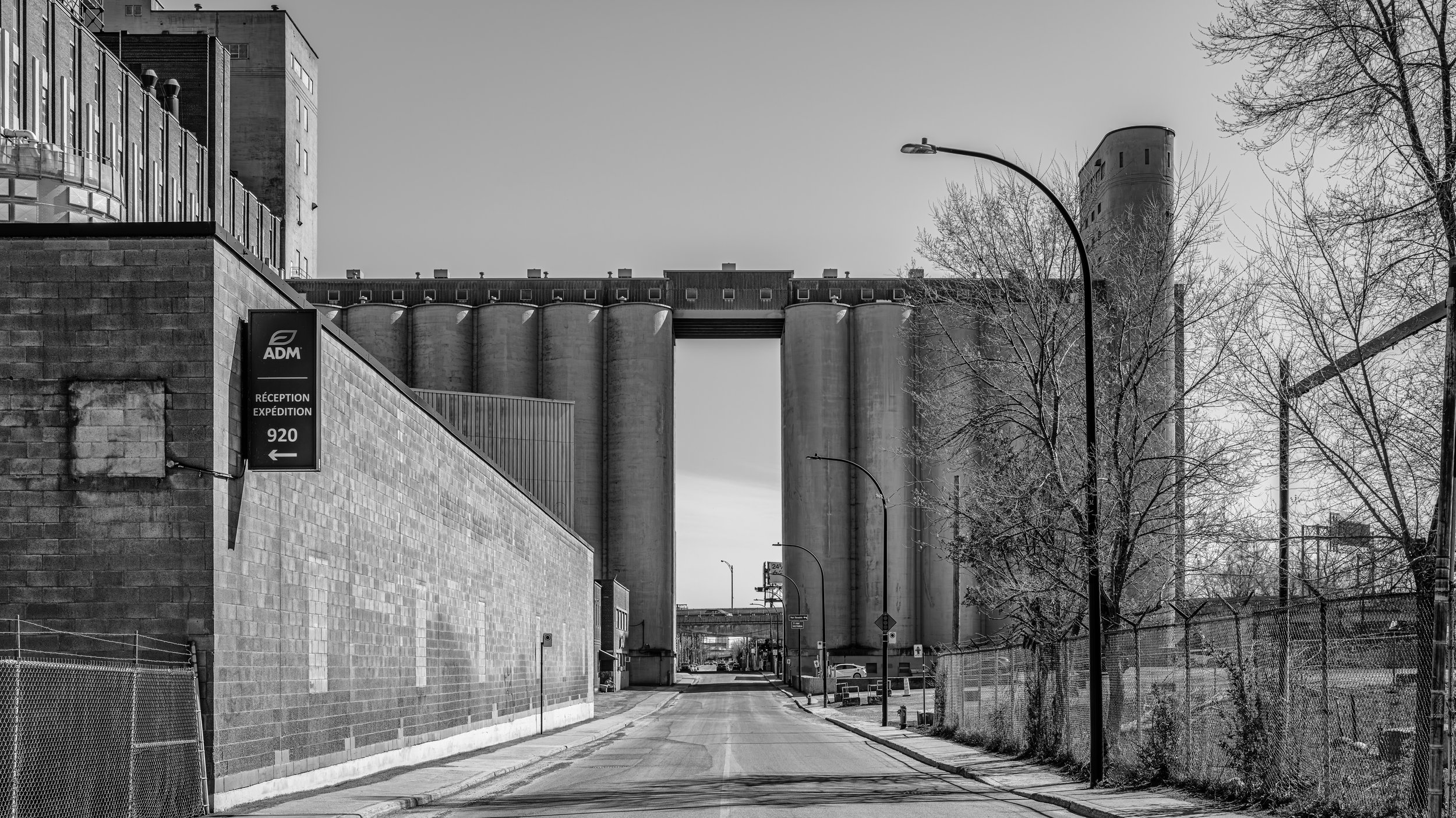 A black and white photo of a city street with industrial buildings, silos, and a brick wall. Bare trees line the sidewalk, and street lamps are visible. A sign on the wall indicates a reception area for a company named ADM with an arrow pointing left