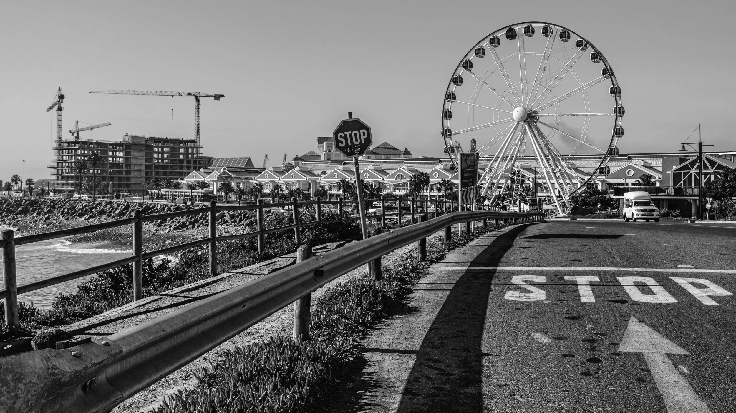 A black and white photo of an amusement park with a large Ferris wheel, a stop sign, and a road with a stop painted on it. There are buildings and construction cranes in the background. Cape Town, South Africa.
