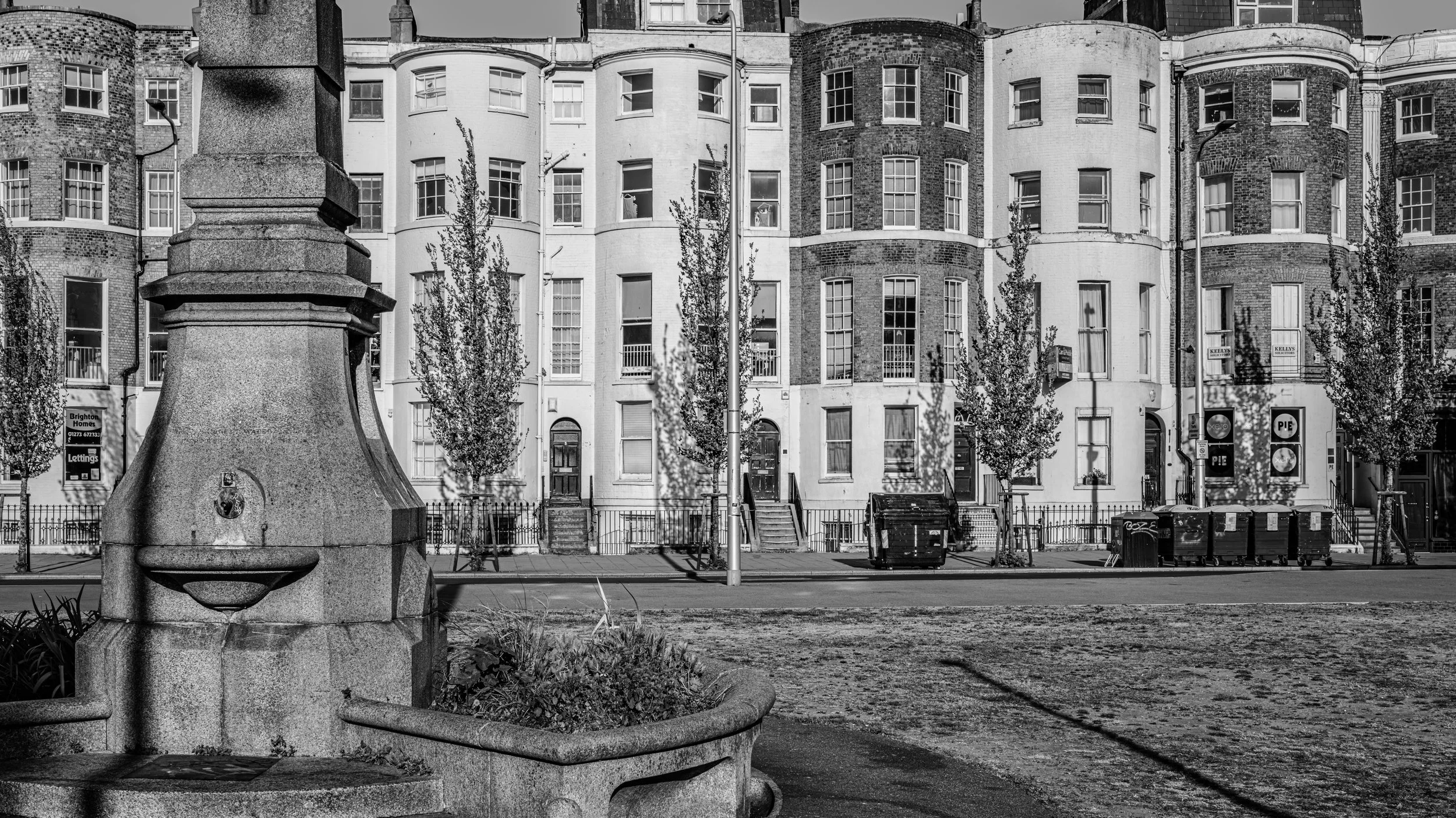 Black and white photo of a city street scene with a stone water fountain in the foreground, a row of multi-story brick and white buildings in the background, trees lining the sidewalk, and trash bins and benches along the street.