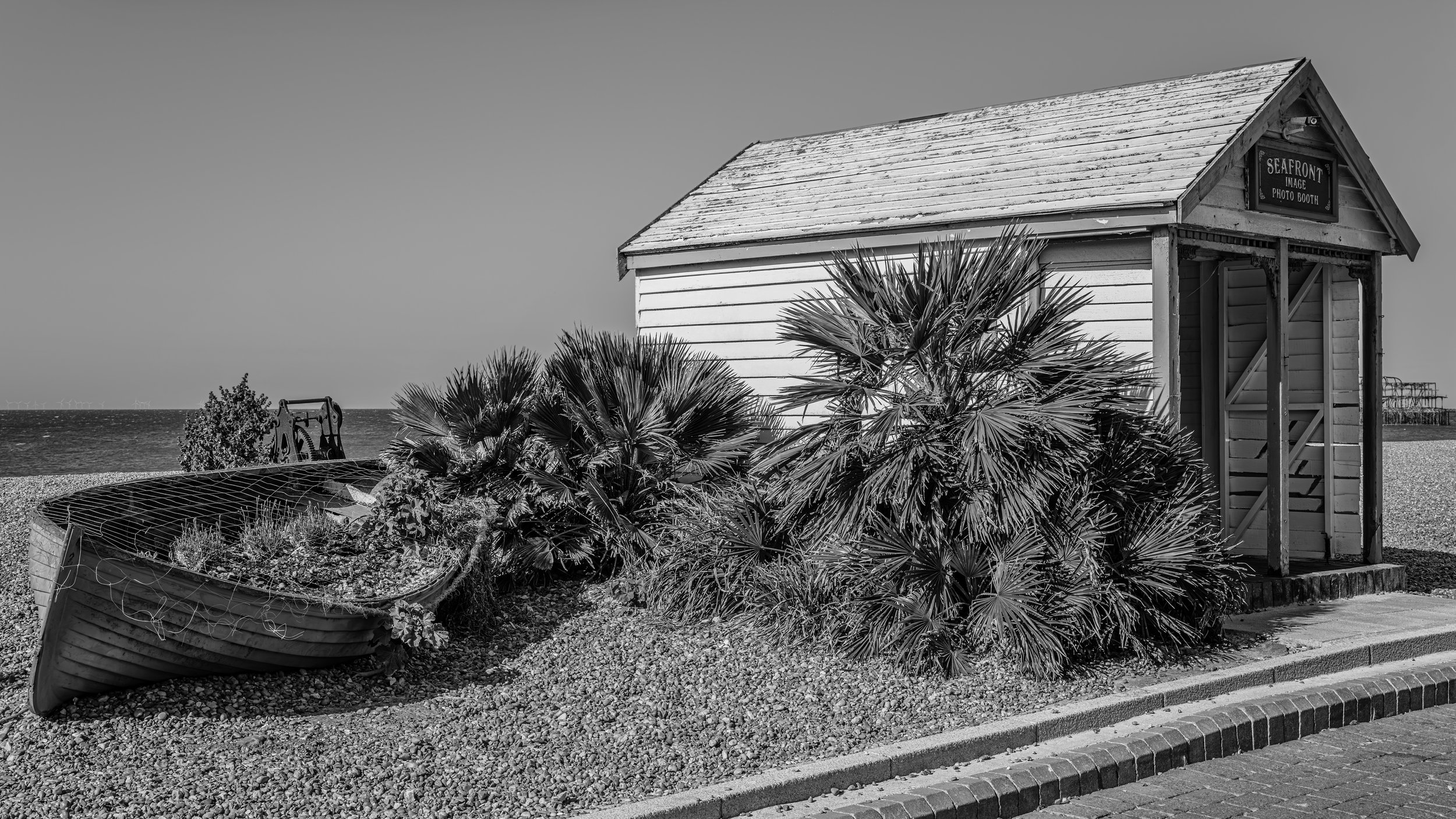 Black and white photo of a small seaside wooden building with a sign reading 'Seafront Image Photo Booth,' surrounded by palm plants, with a body of water and wind turbines in the distance.