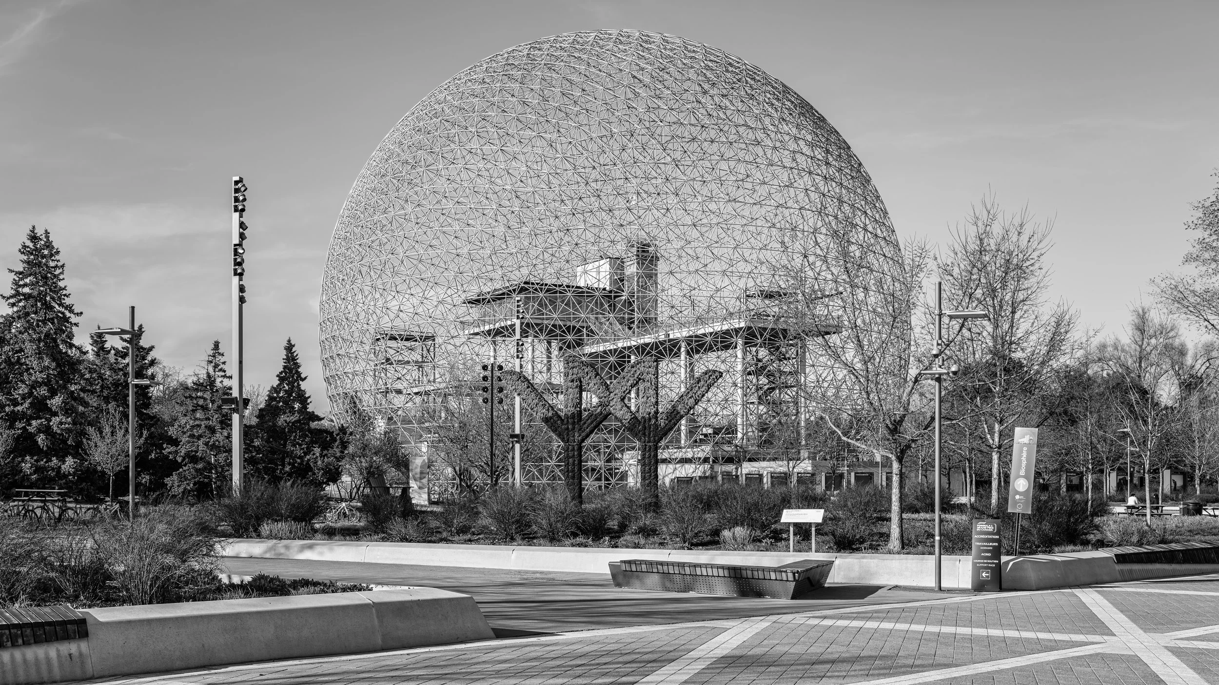 A large geodesic dome structure surrounded by trees and park features, with pathways, benches, and signage in a park setting.