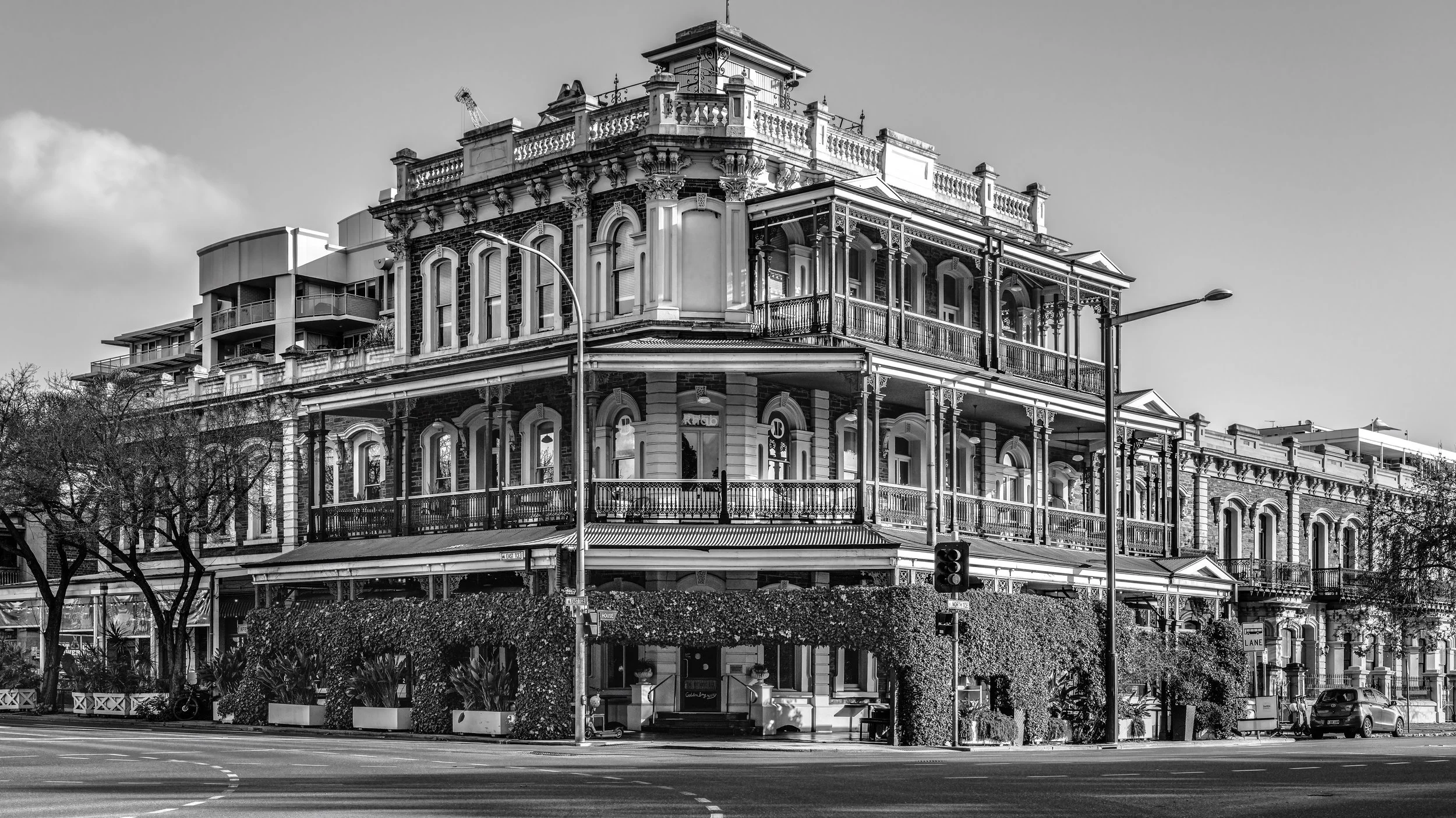 A historic multi-story building with intricate architectural details, including decorative columns, balconies, and a rooftop railing, situated on a city street corner with trees, streetlights, and vehicles in the foreground.