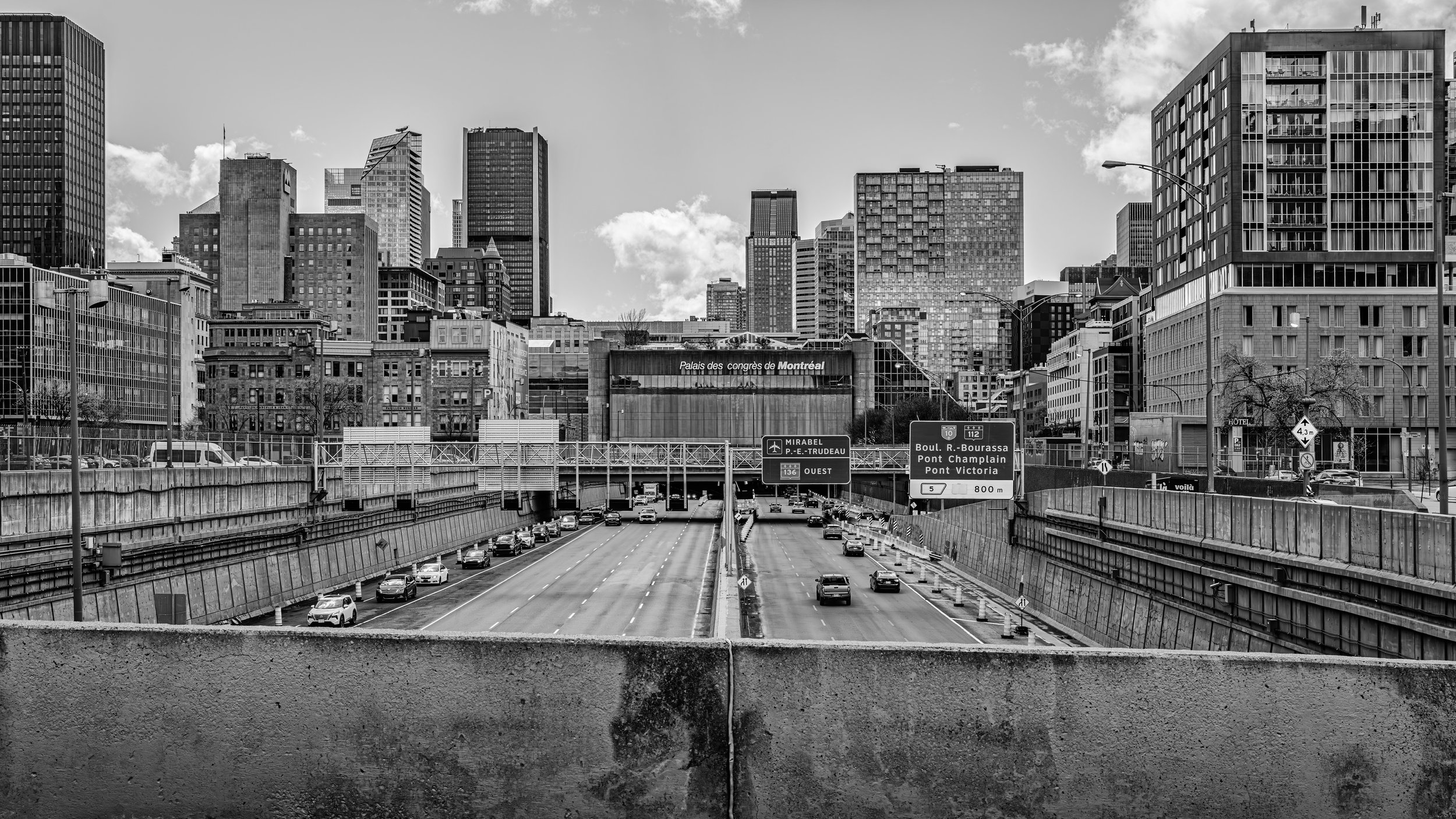 A black and white photo of a city skyline with tall buildings. There is a highway with cars in the foreground and signs indicating directions and distances.