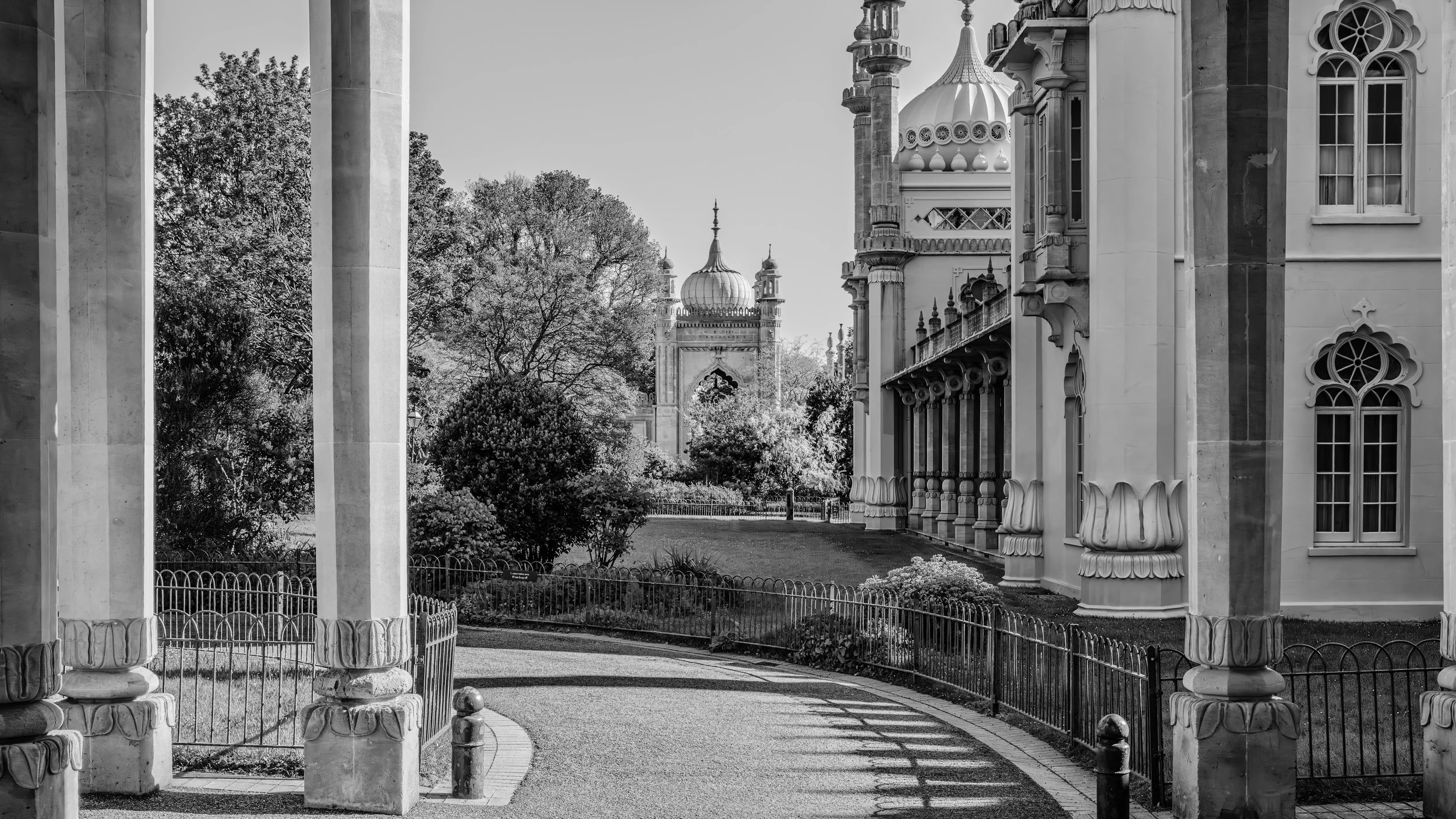 Black and white photo of an ornate palace with domes and towers in the background, viewed through a colonnade and garden with trees and a pathway in the foreground.