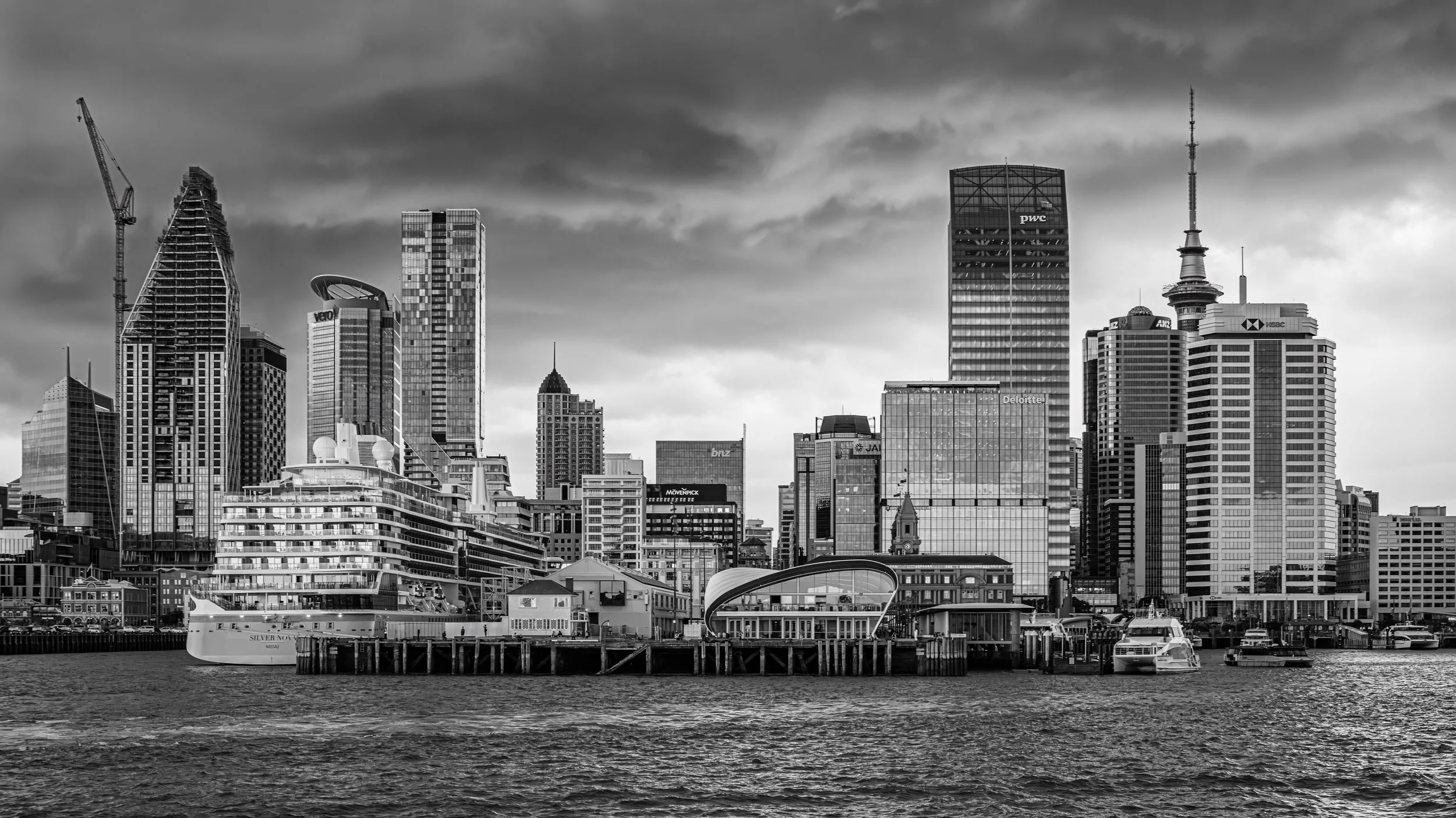 Black and white city skyline with skyscrapers, a waterfront with boats and a pier in the foreground.