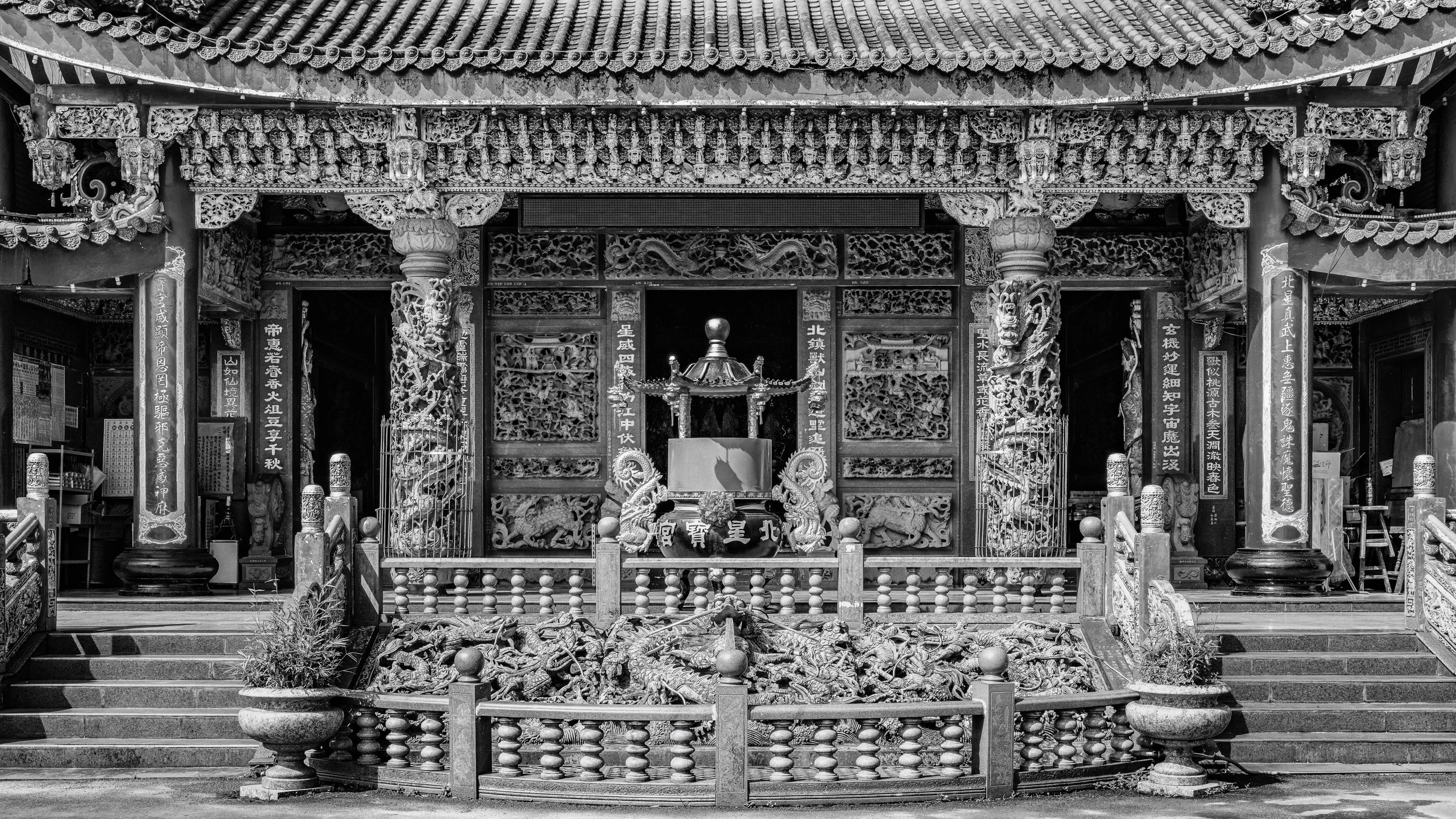 Black and white photo of a traditional Asian temple entrance with intricate carvings, dragon sculptures, columns, banners with Chinese characters, and steps leading up to a doorway.