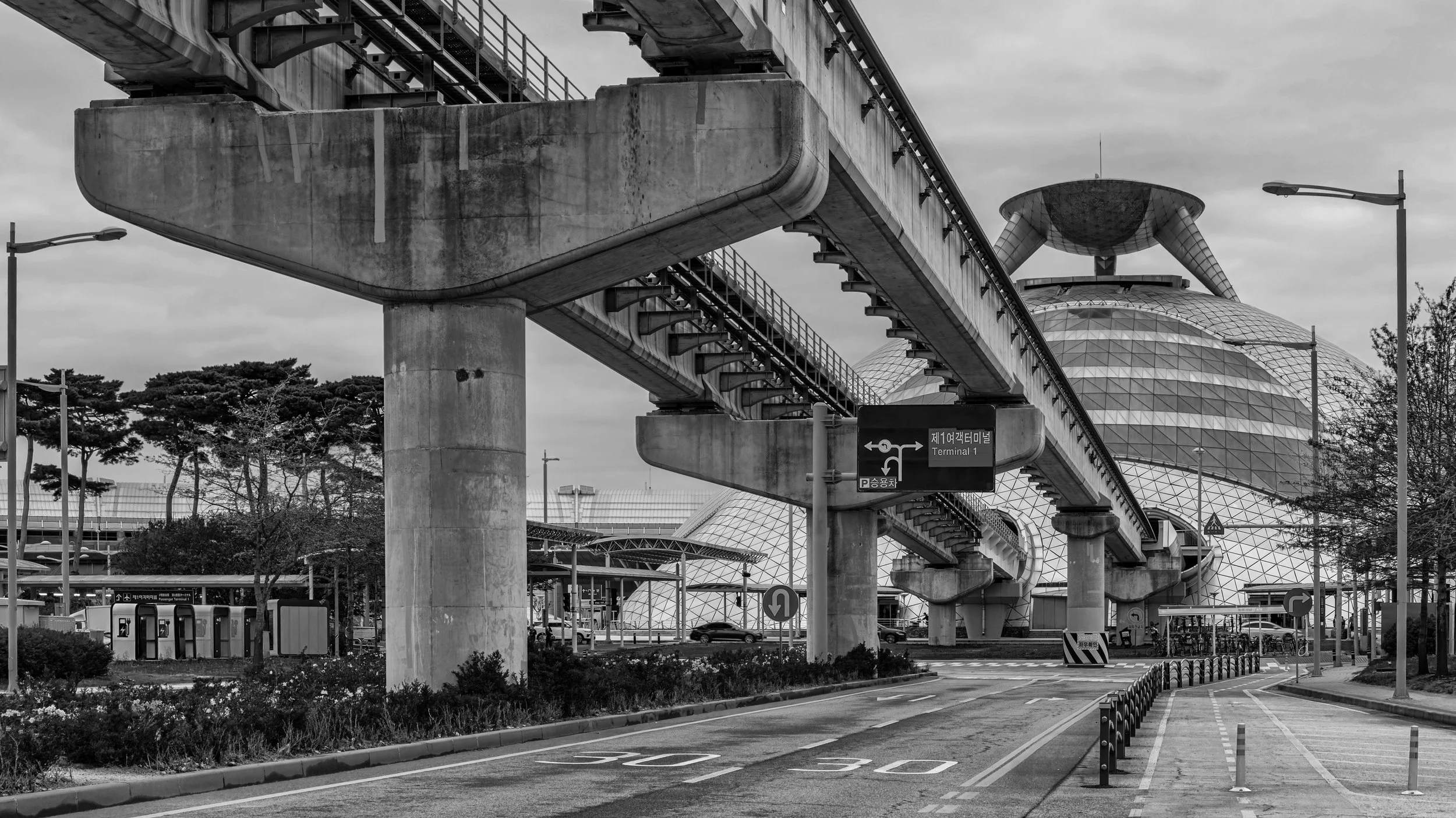 Black and white photograph of an elevated train track over a busy urban area with futuristic buildings, street lamps, and traffic signs, including a sign indicating 'Terminal 1' and parking directions.
