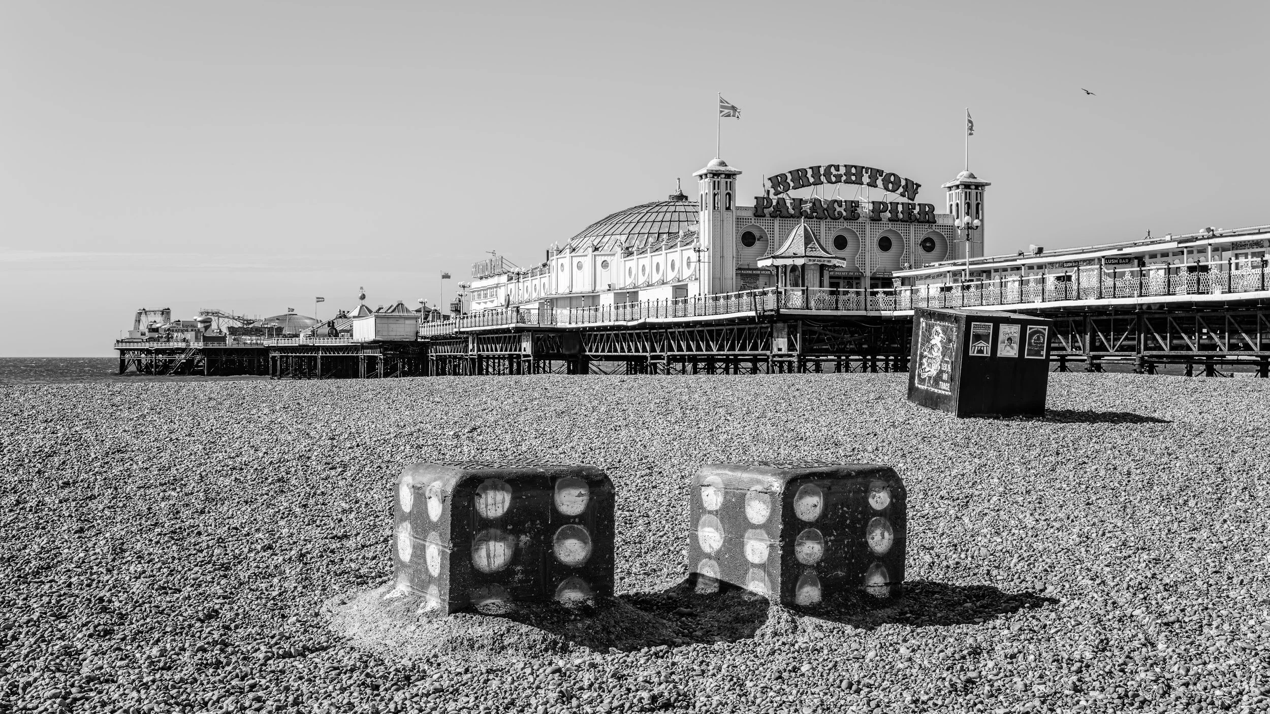 Black and white photo of Brighton Palace Pier with a pebble beach in the foreground, large advertising sign, and a clear sky.