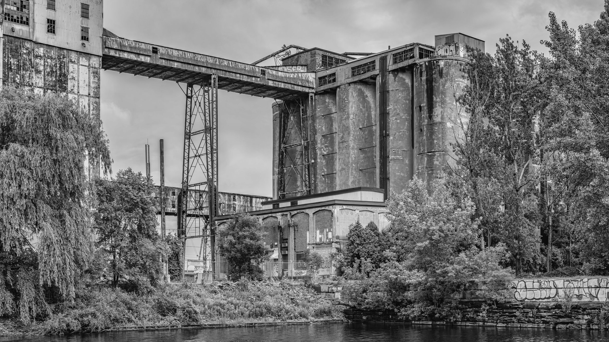 Abandoned industrial building with a large bridge structure and trees, located beside a body of water in black and white.