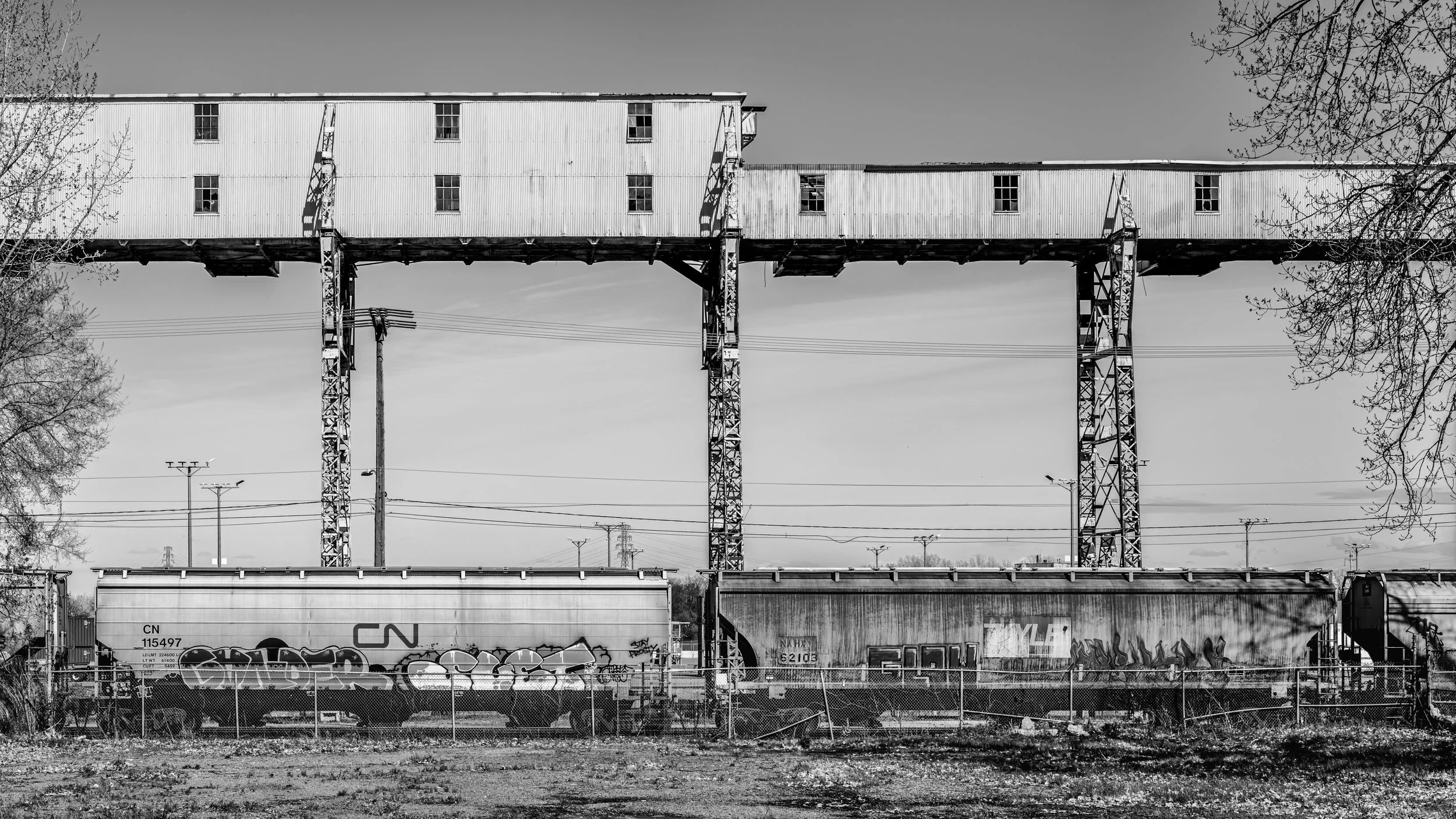 Black and white photo of an old elevated building on steel supports above train cars. Trees frame the sides, and power lines are visible in the background.