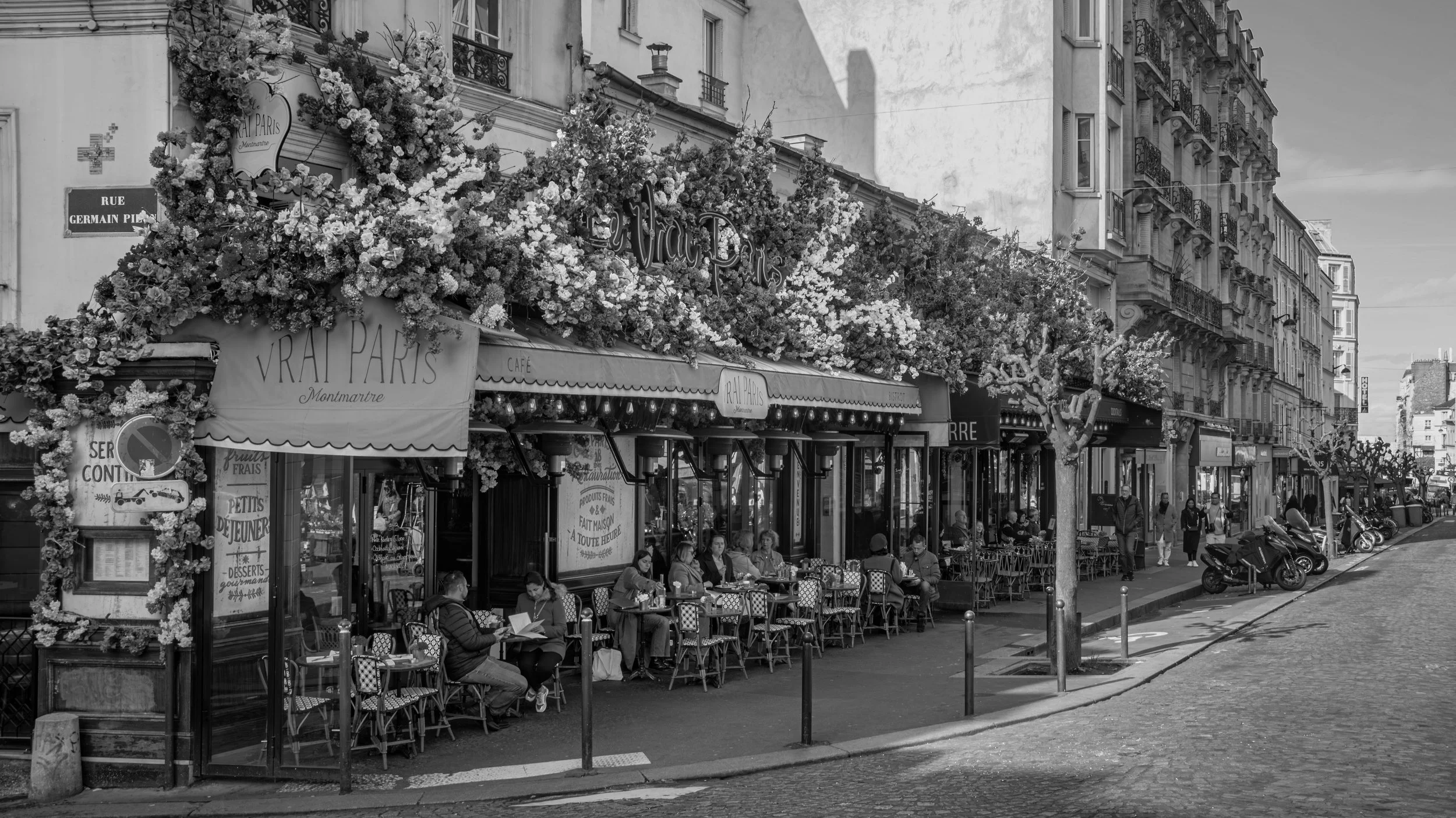 Street scene in Paris with a bustling sidewalk cafe covered in flowers, people sitting at tables, and buildings with balconies lining the street.