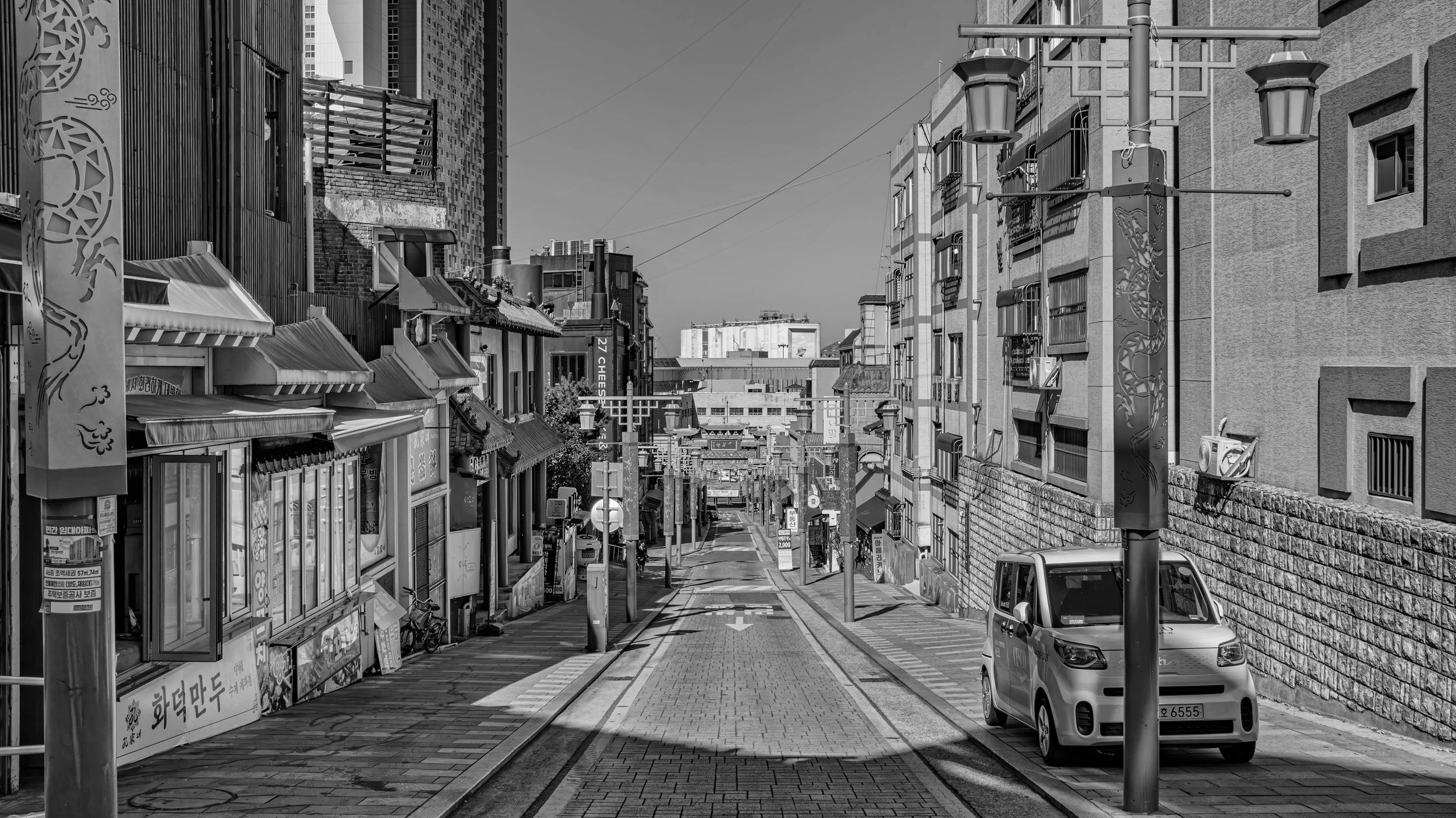 A black and white photo of a city street with buildings on both sides, parked cars, street lamps, and shops with Korean signs. The street has a slight incline, leading towards taller buildings in the background.